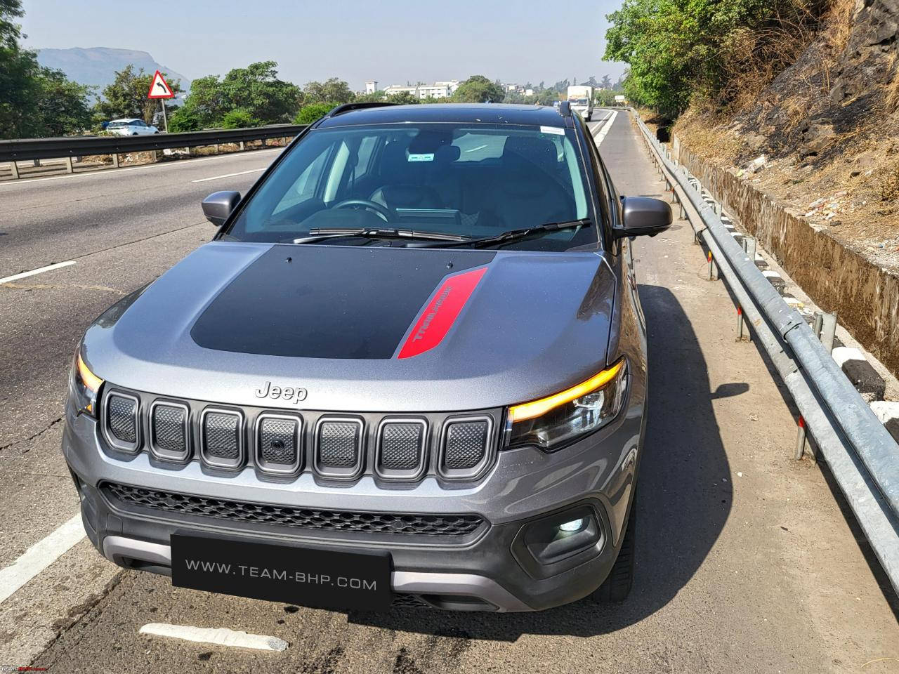 灰色Jeep Compass，帶紅色條紋，停在陽光明媚的高速公路旁，靠近綠樹和山丘。