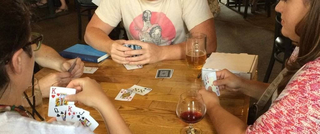 精於牌技的玩家進行Écarté遊戲 壁紙 — 三個人圍著木桌玩牌，桌上有飲品和一本書，在舒適的室內環境中。