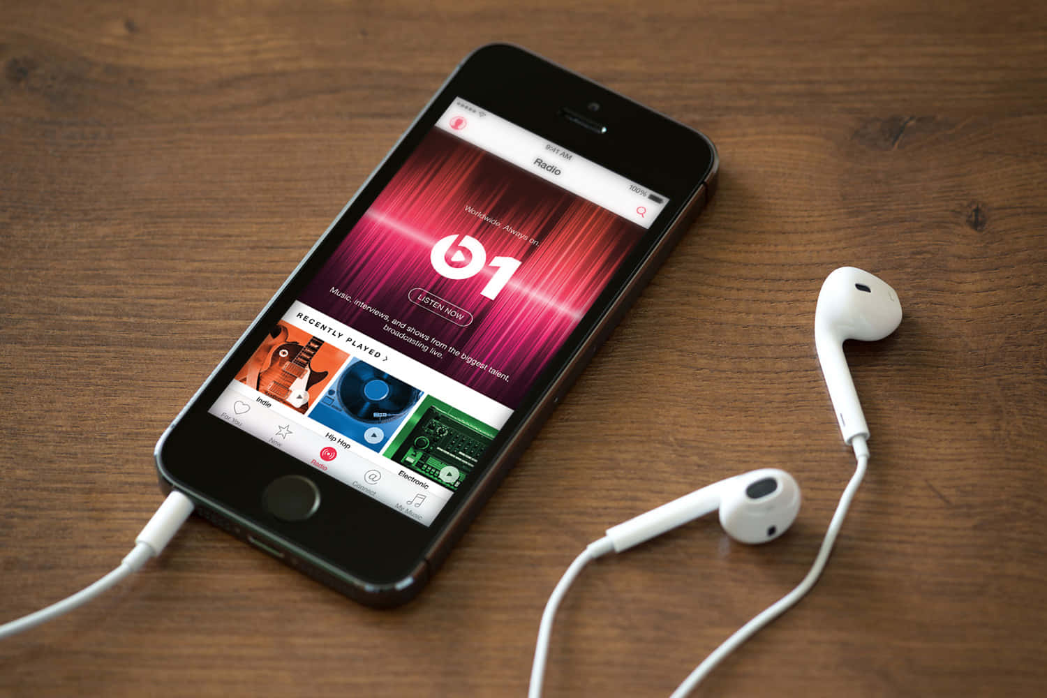 精緻 Apple 智能手機，深色背景襯托 壁紙 — 一部顯示 Beats 1 收音機應用程式的智能手機放在木桌上，旁邊有白色有線耳機。