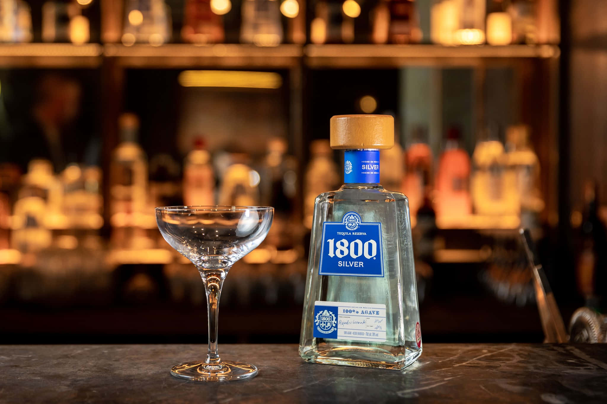 精緻1800龍舌蘭酒Reserva Silver擺放於吧檯 壁紙 — 一個透明嘅1800 Silver龍舌蘭酒樽，木蓋，旁邊係一個空嘅coupe杯，喺黑暗嘅吧枱上，背景有溫暖模糊嘅層架。