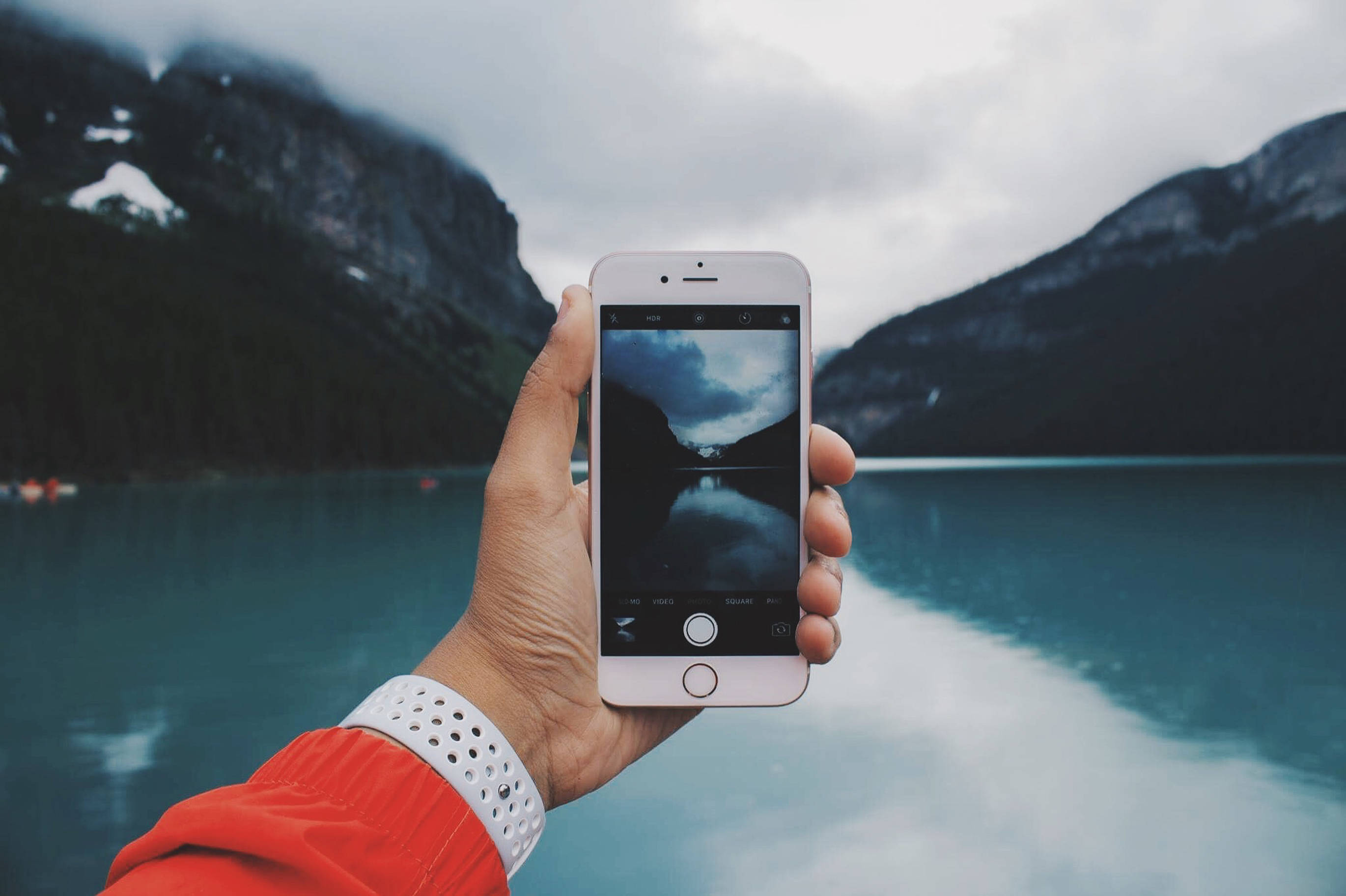 精緻iPhone 8配簡約優雅背景 壁紙 — 一隻手拿著智能手機拍攝一個霧氣繚繞的山湖，有積雪的山峰和寧靜的碧綠湖水。