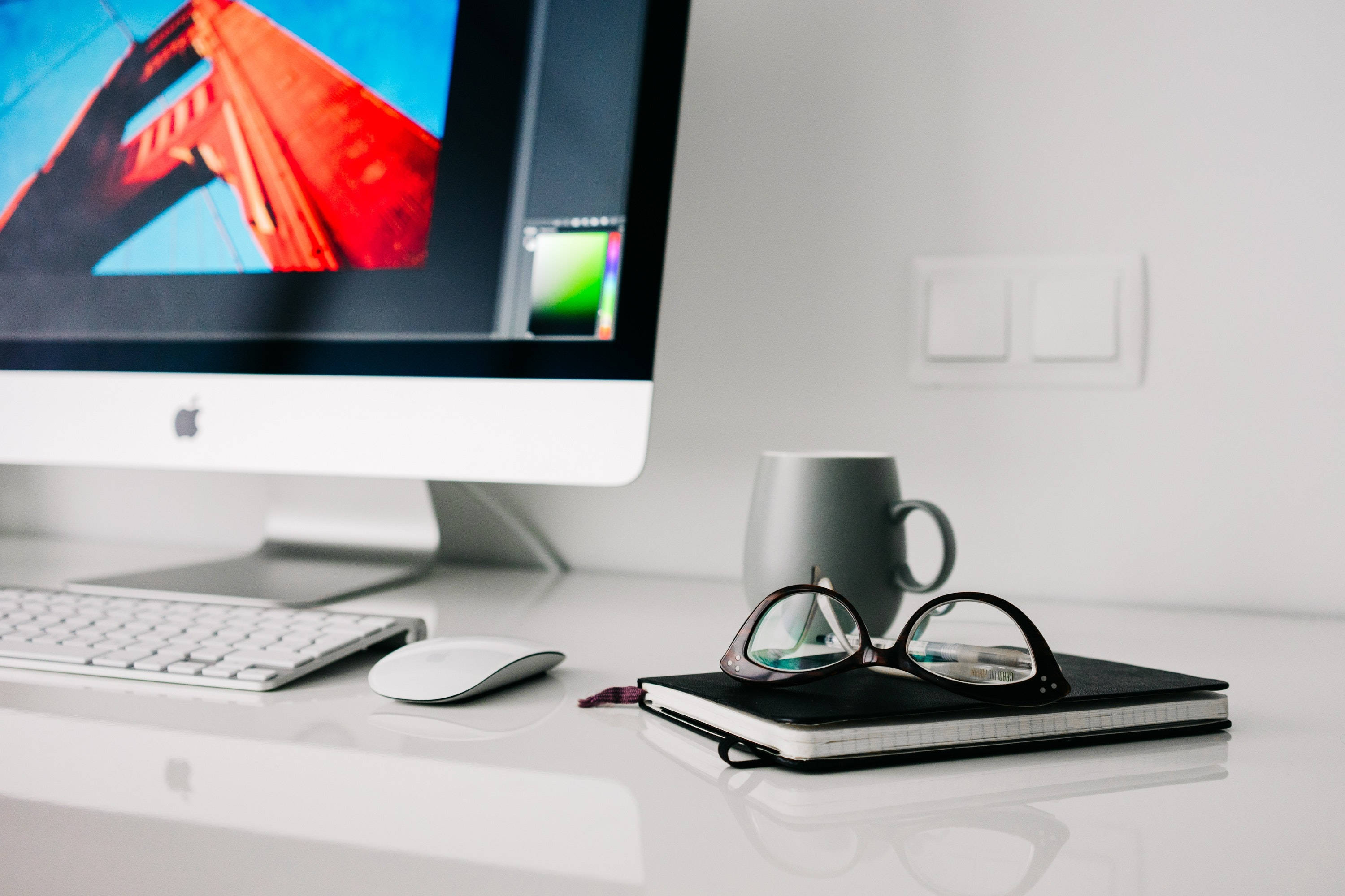 精緻Mac工作區佈置於潔淨木桌之上 壁紙 — 簡約白色桌面設置，iMac 顯示紅色橋樑壁紙，灰色馬克杯和眼鏡放在筆記本上。