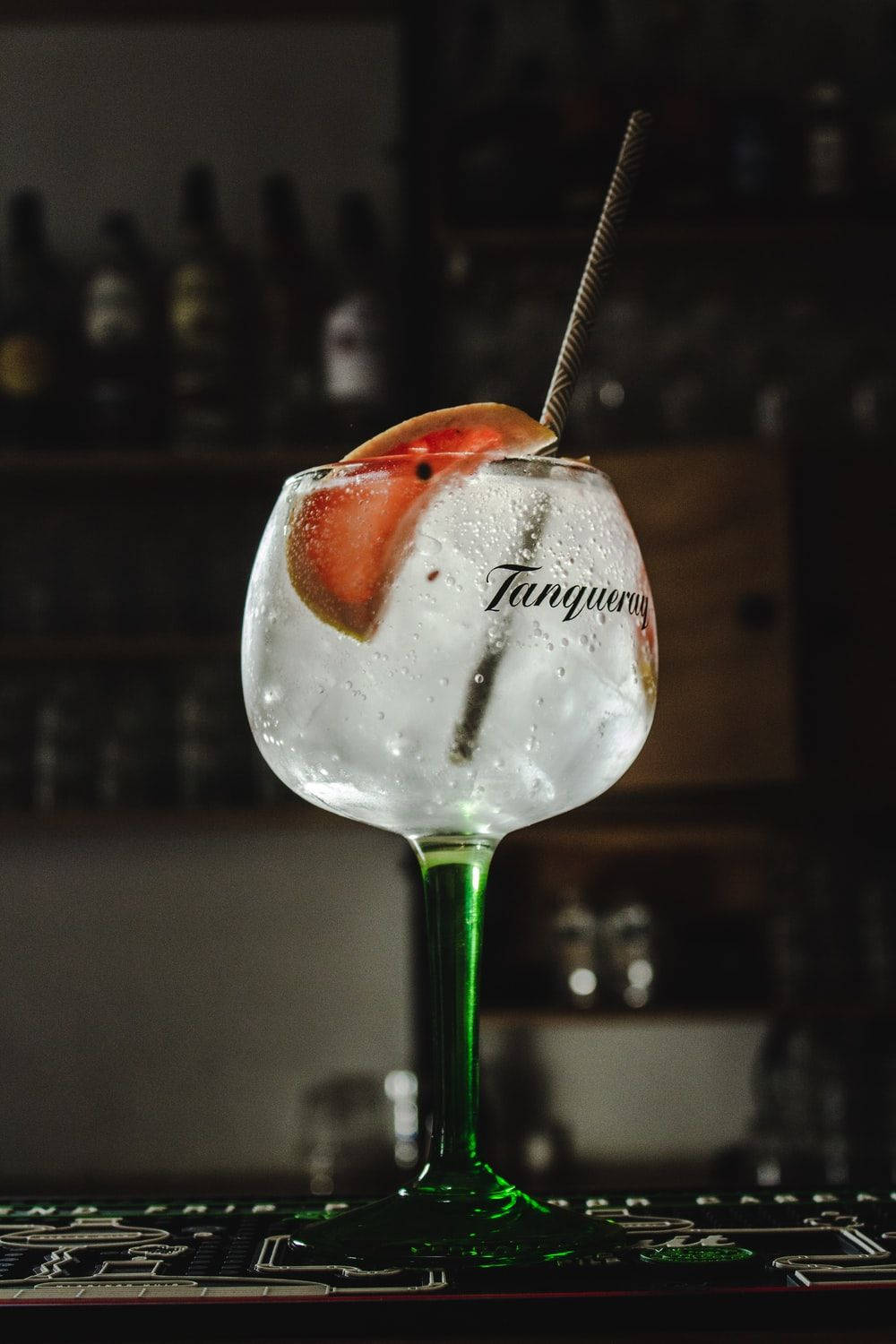 精緻Tanqueray金酒盛於高腳杯 壁紙 — 一杯冰鎮氈酒湯力水放在品牌玻璃杯中，飾以西柚片和飲管，置於酒吧上，背後有模糊的酒瓶。
