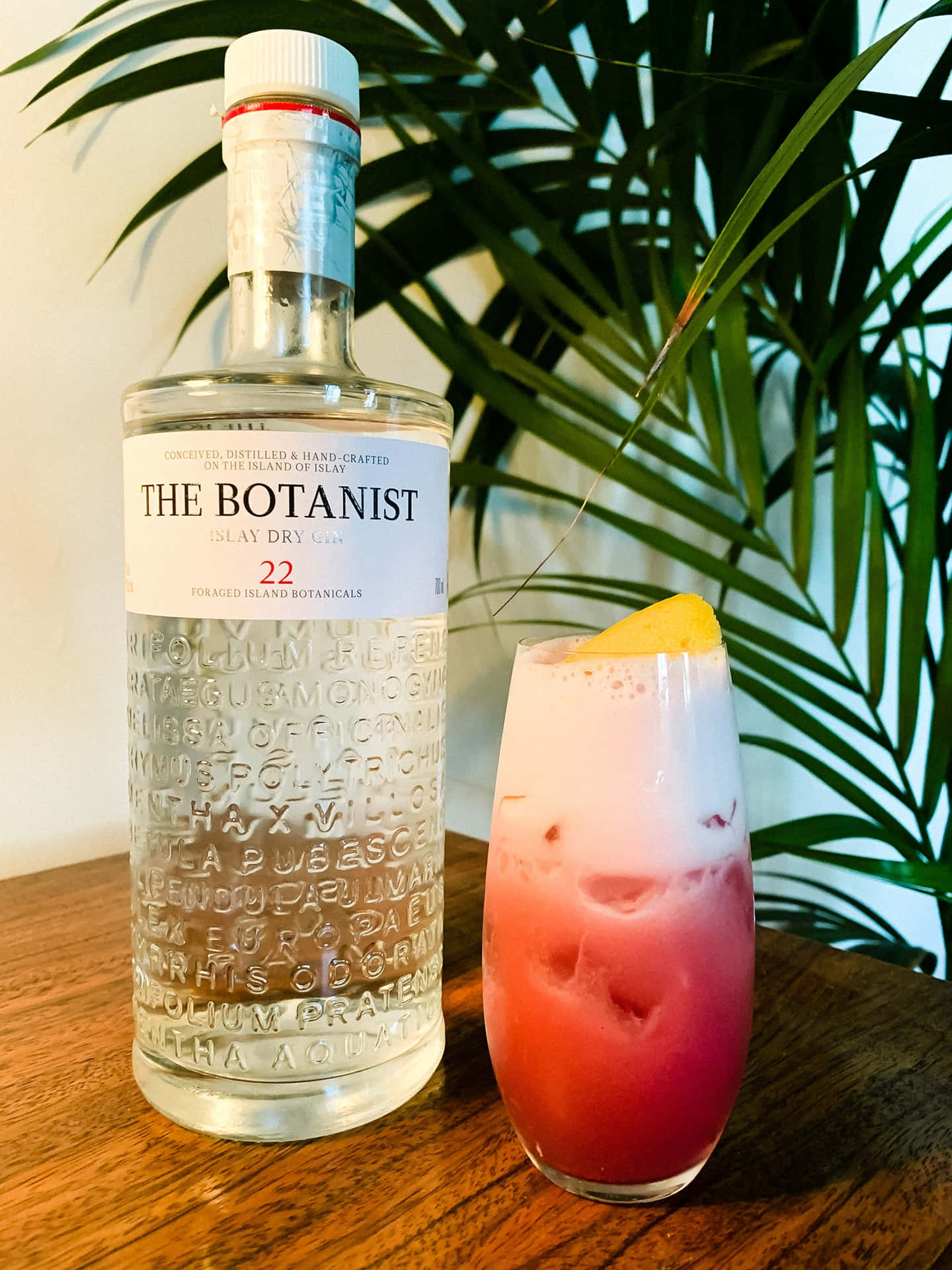 Botanist 氈酒瓶和粉紅色雞尾酒配檸檬，木桌上，棕櫚葉旁。