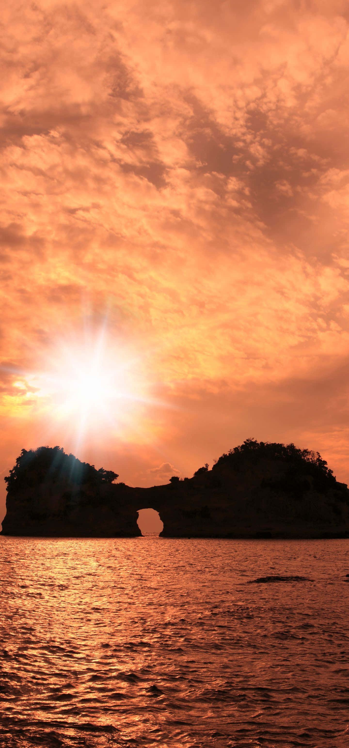 精緻灰調壁紙紋理 壁紙 — 燦爛的夕陽在平靜的海面上投射出熾熱的橙色光芒，在戲劇性的天空下照亮了天然岩石拱門的輪廓。