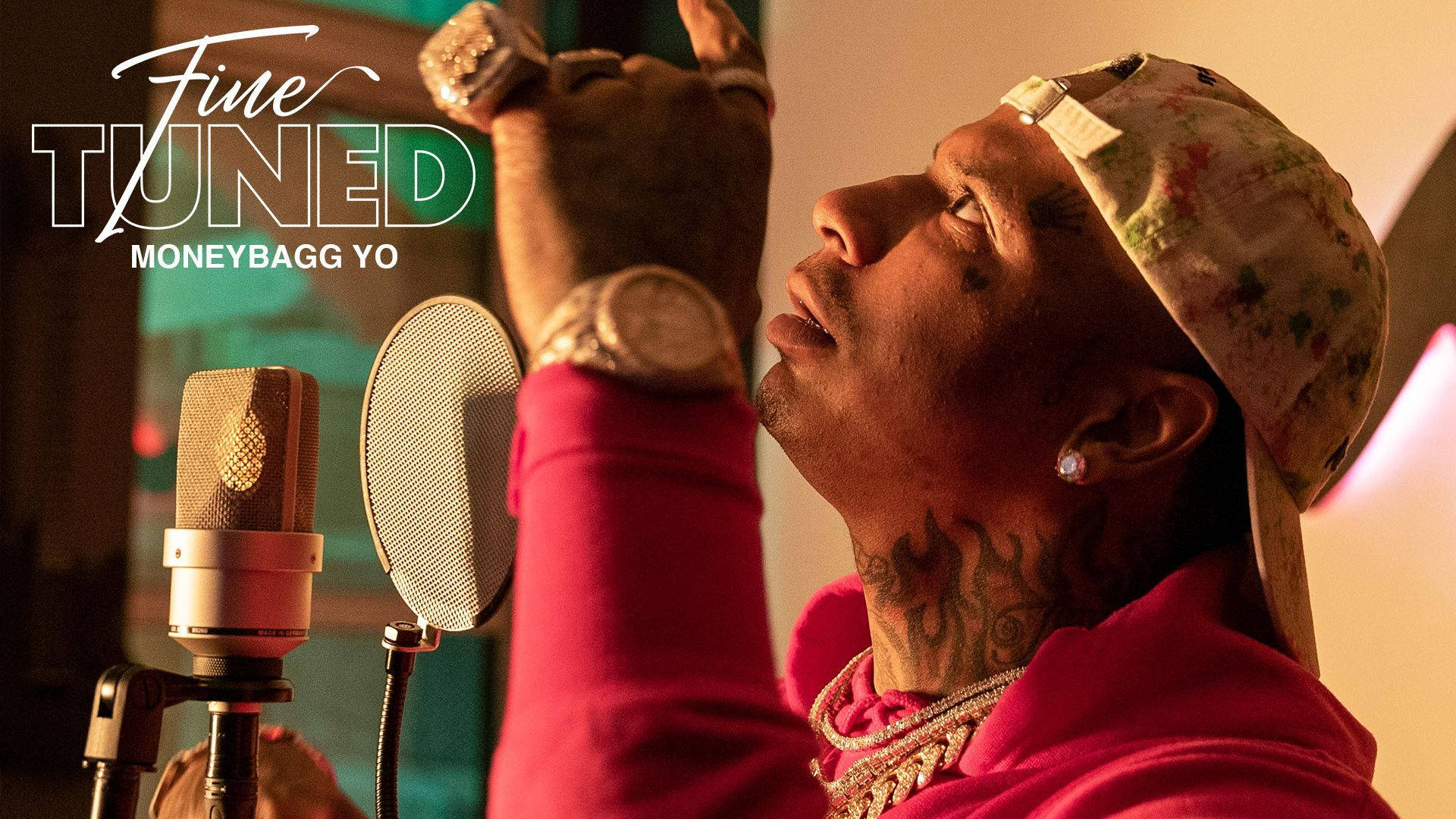 精緻版 Moneybagg Yo 壁紙 — 饒舌歌手 Moneybagg Yo 身穿粉紅色連帽衫和花卉圖案帽子，在溫暖照明的錄音室中錄音時向上做手勢。