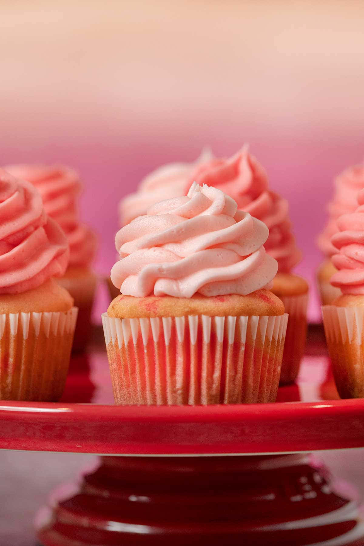 Pink Cupcakes 桌布 壁紙 — 精緻的粉紅色糖霜紙杯蛋糕放在白色襯紙中，優雅地擺放在光澤的紅色蛋糕架上，背景柔和漸變。