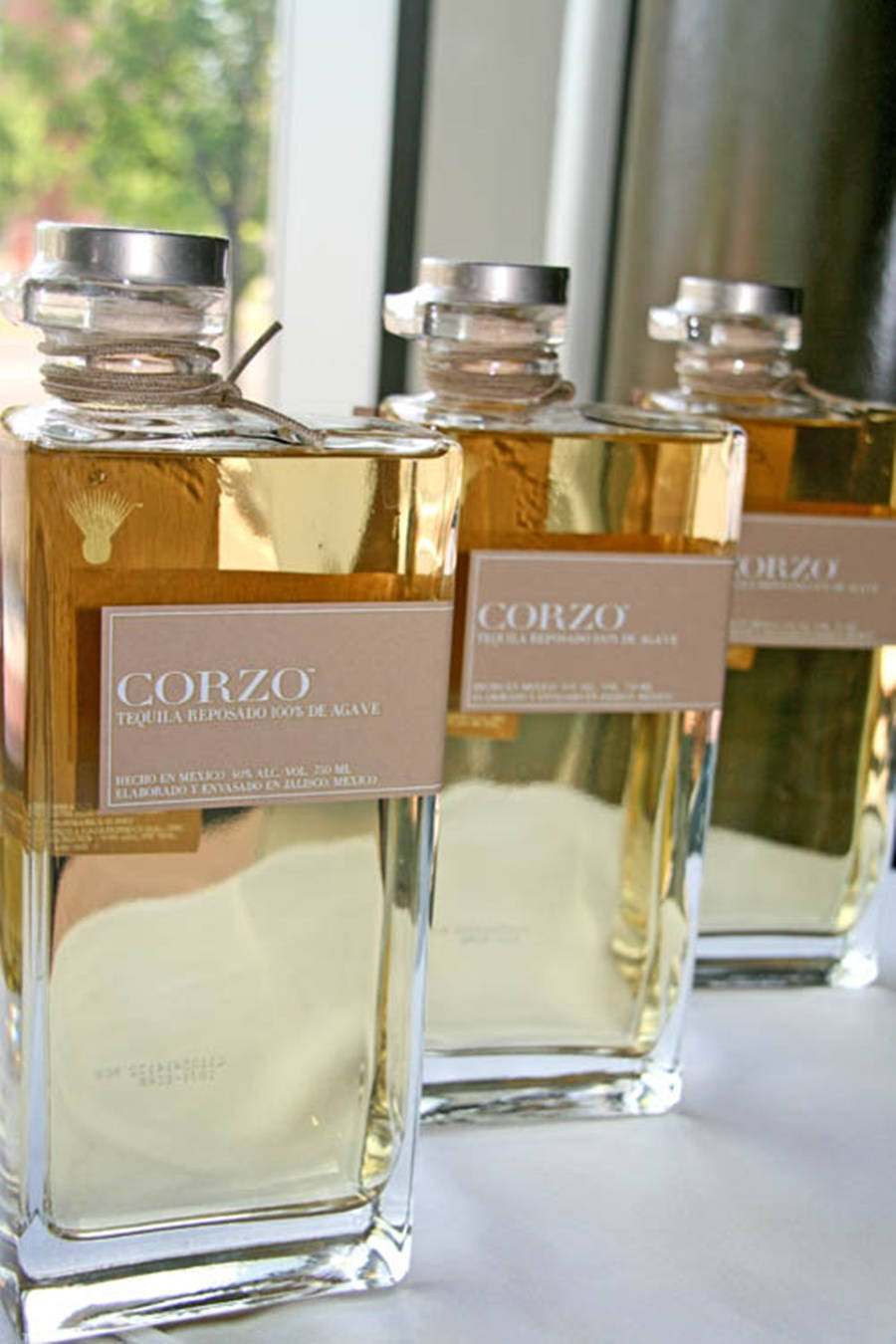 三瓶Corzo龍舌蘭酒，裝有金色液體，放在白色表面靠近窗邊。