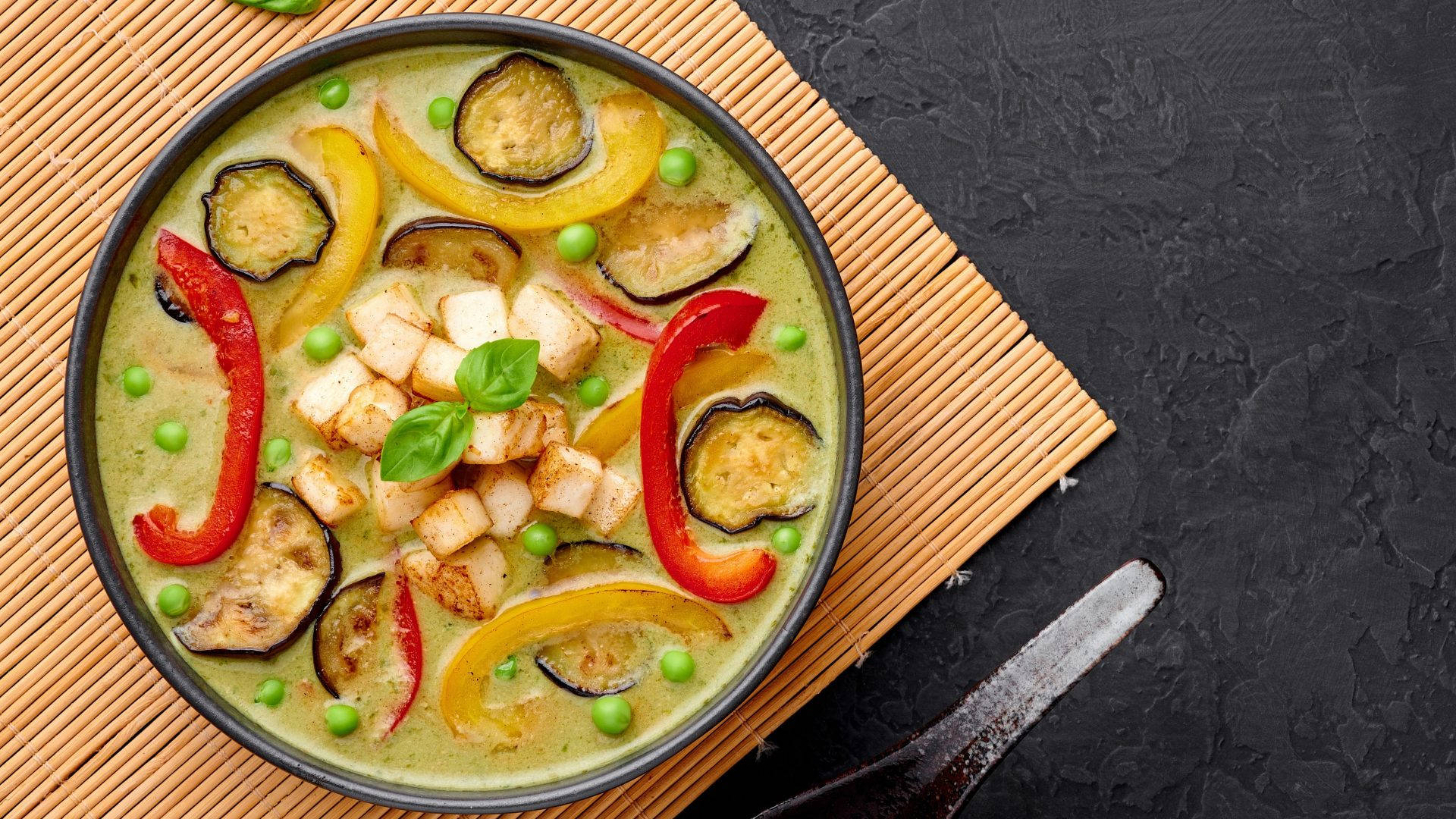 黑色碗中的青咖喱，配有豆腐、茄子和甜椒，置於竹蓆上。