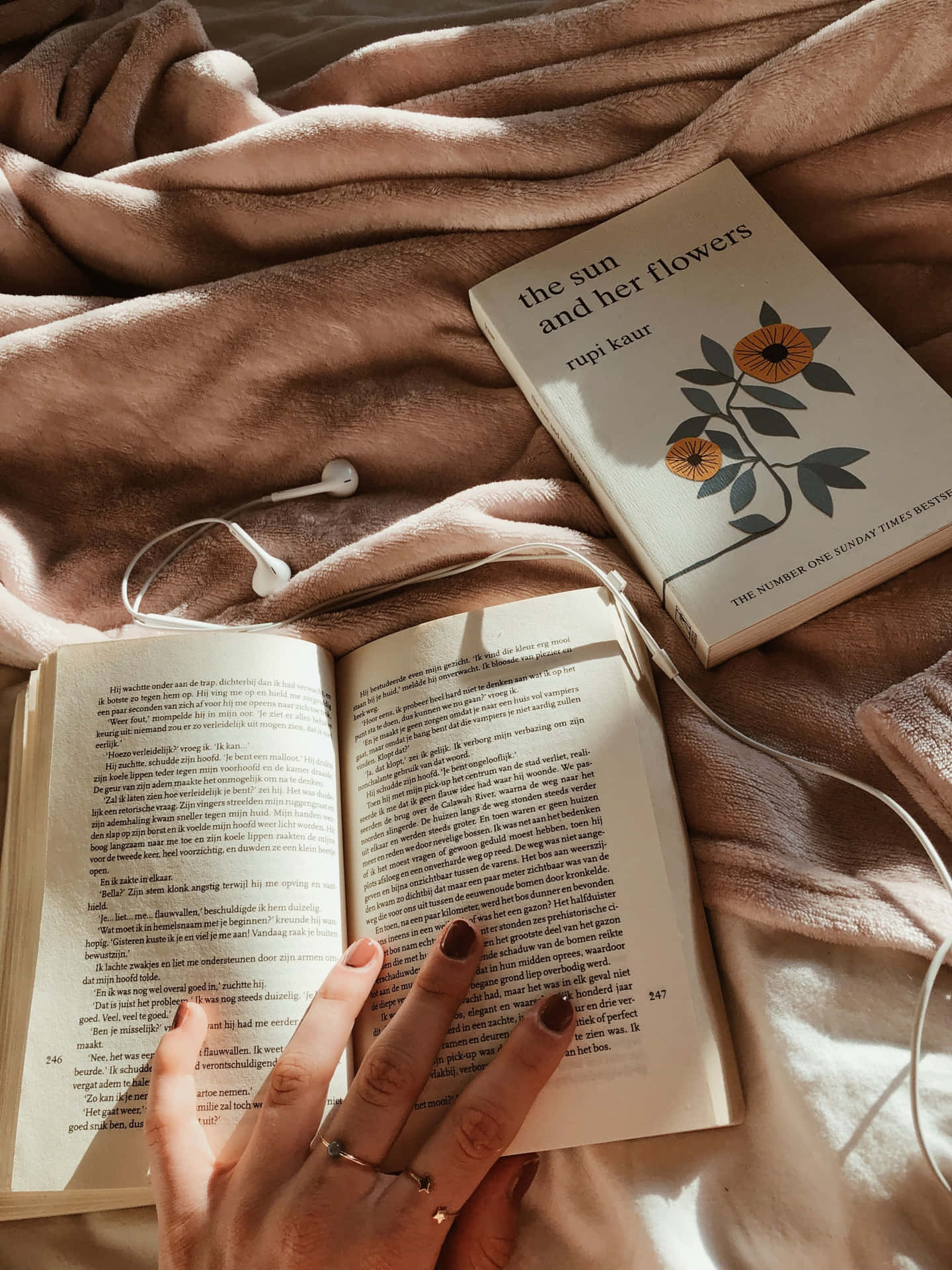 精緻美學藝術書籍 壁紙 — 一隻手放在一本名為《the sun and her flowers》的打開書本上，作者是rupi kaur，旁邊有一本合上的書和白色耳塞，放在柔軟的粉紅色毯子上。
