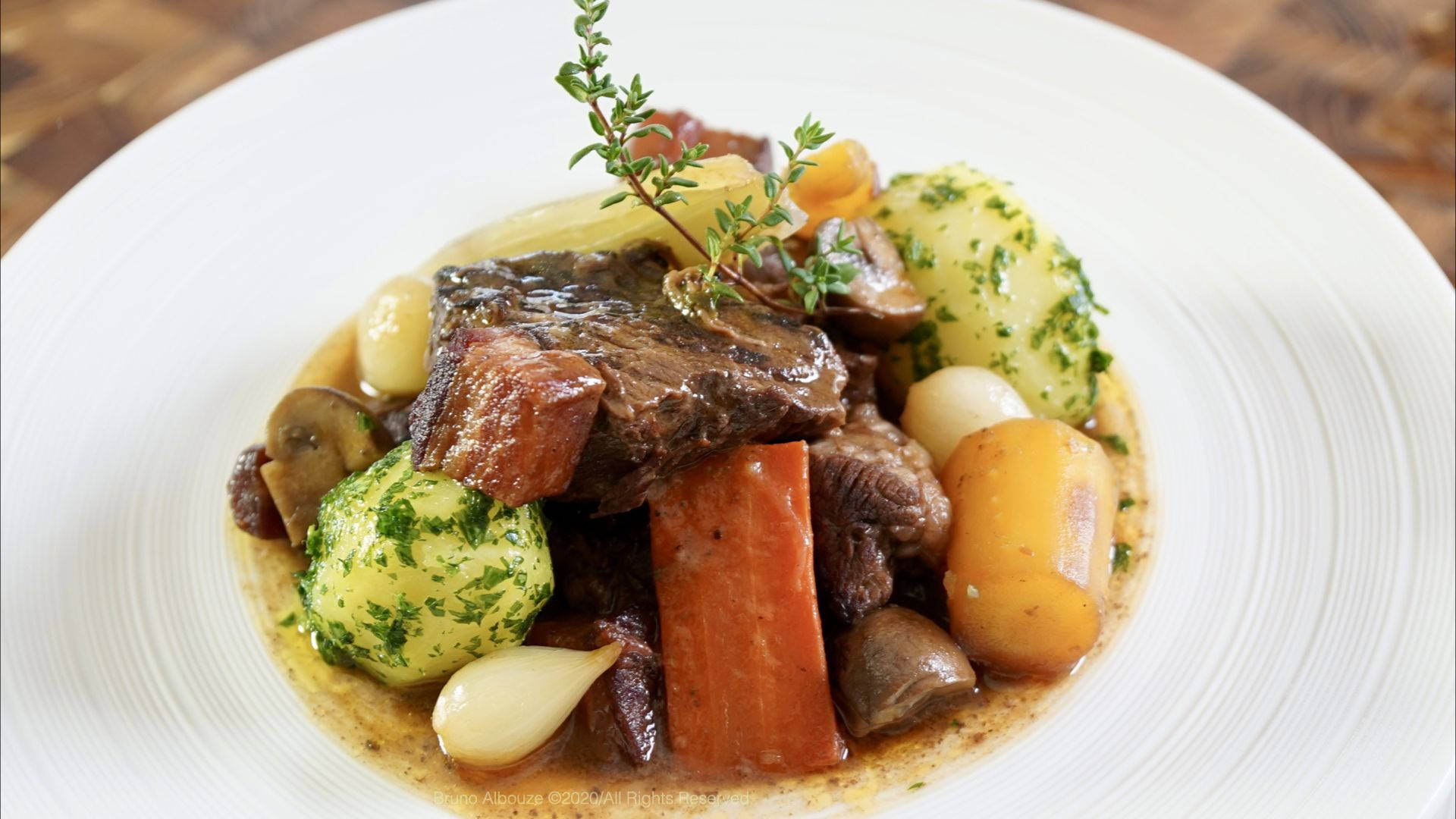 精緻裝飾的牛肉布丁 壁紙 — 豐富的燉牛肉，搭配紅蘿蔔、馬鈴薯和香草，盛載於白色碟子上，背景為質樸的木質桌面。