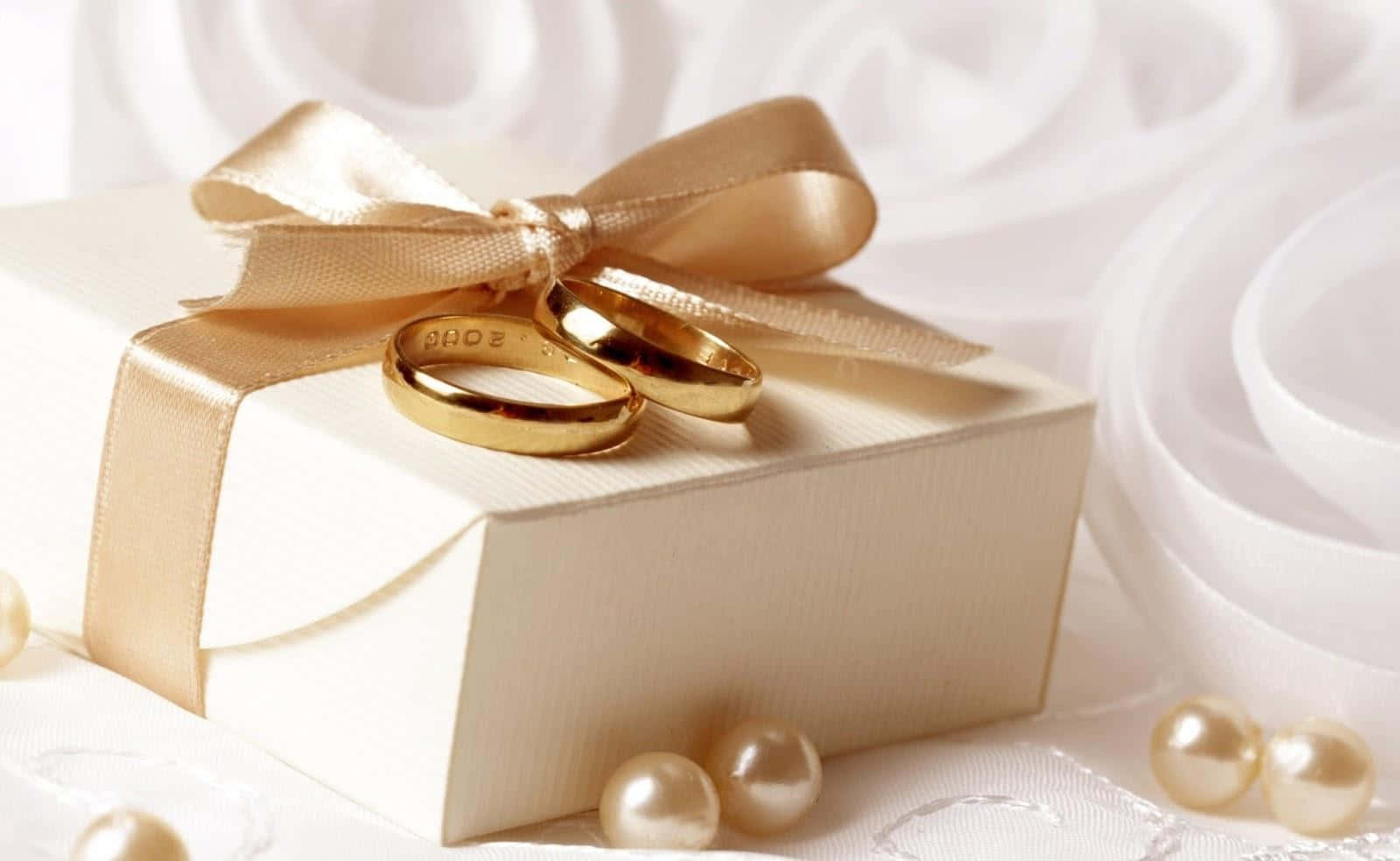 精緻鑽石戒指展示 壁紙 — 兩枚金色結婚戒指放在一個用金色絲帶綁著的奶油色禮物盒上，周圍環繞著珍珠和白色布料。