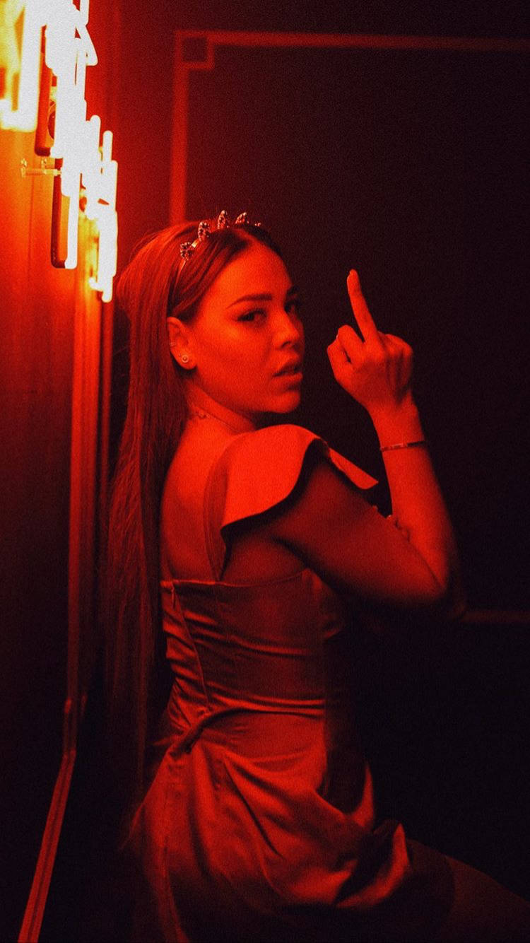《精英》丹娜中指動作 壁紙 — 一位身穿紅色連衣裙的女性在昏暗的房間裡站在霓虹燈下做出挑釁的手勢。