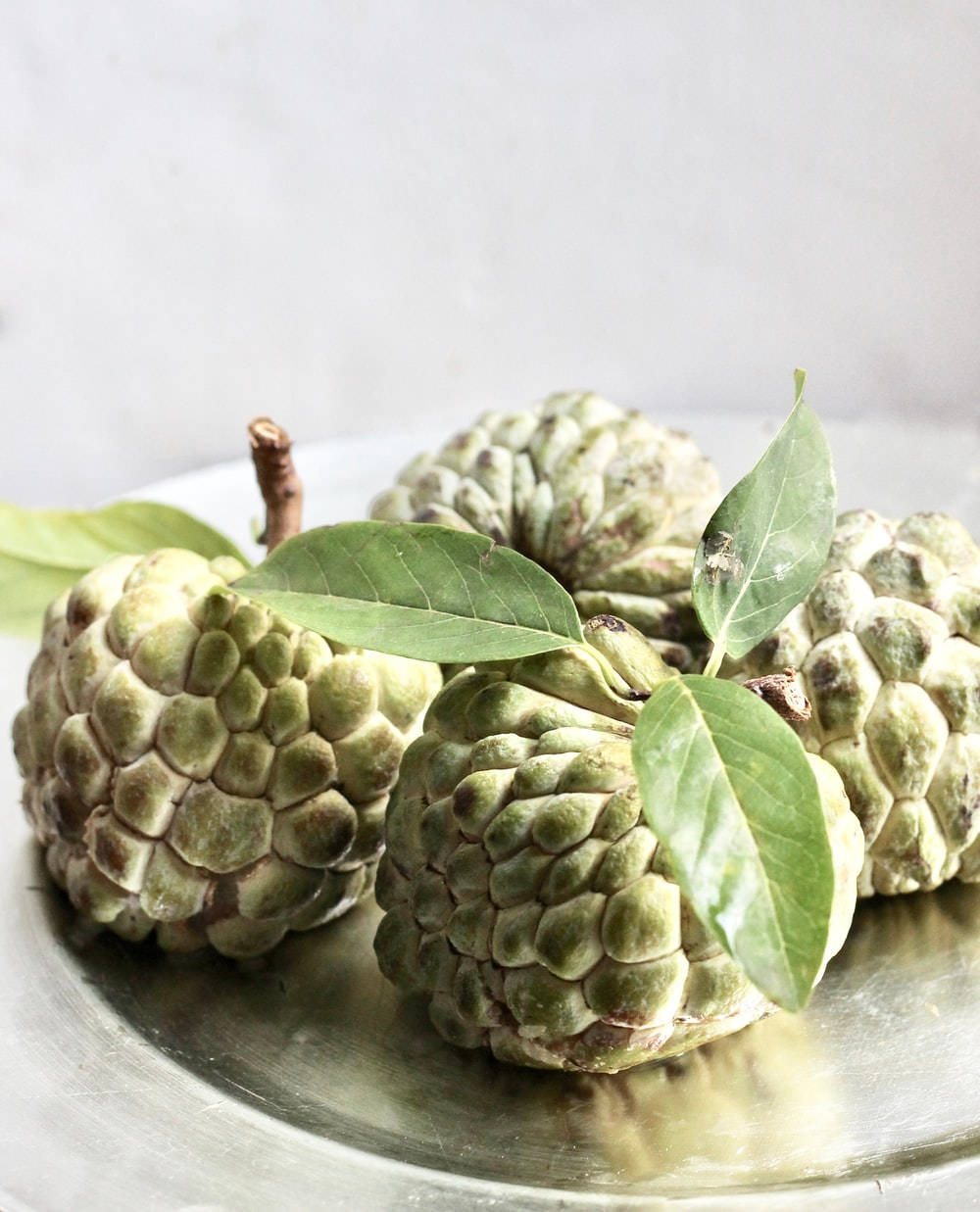 糖蘋果甜品果實 壁紙 — 新鮮綠色嘅牛油果（custard apples）連葉放喺反射性銀碟上，背景係柔和白色。