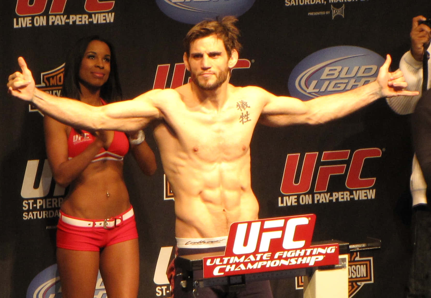 約恩·菲奇UFC稱重儀式擺姿 壁紙 — 一位肌肉發達的UFC拳擊手在品牌背景前，自信地在體重磅上擺姿勢，旁邊有一位微笑的擂台女郎。