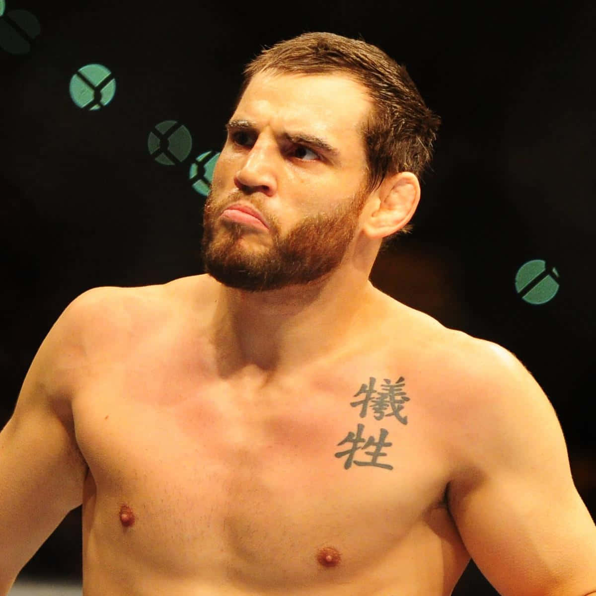 約恩·菲奇於2011年Acer Arena UFC 127賽前 壁紙 — 一個肌肉發達、留鬍子的戰士，表情嚴厲，胸前有紋身，在昏暗的競技場中凝視著。