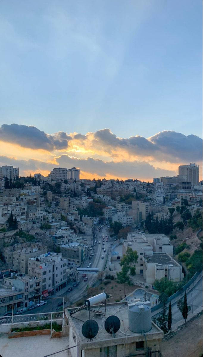 約旦日出 壁紙 — 夕陽將雲彩染成金色，籠罩著一座密集的山丘城市，蜿蜒的道路和屋頂水箱清晰可見。