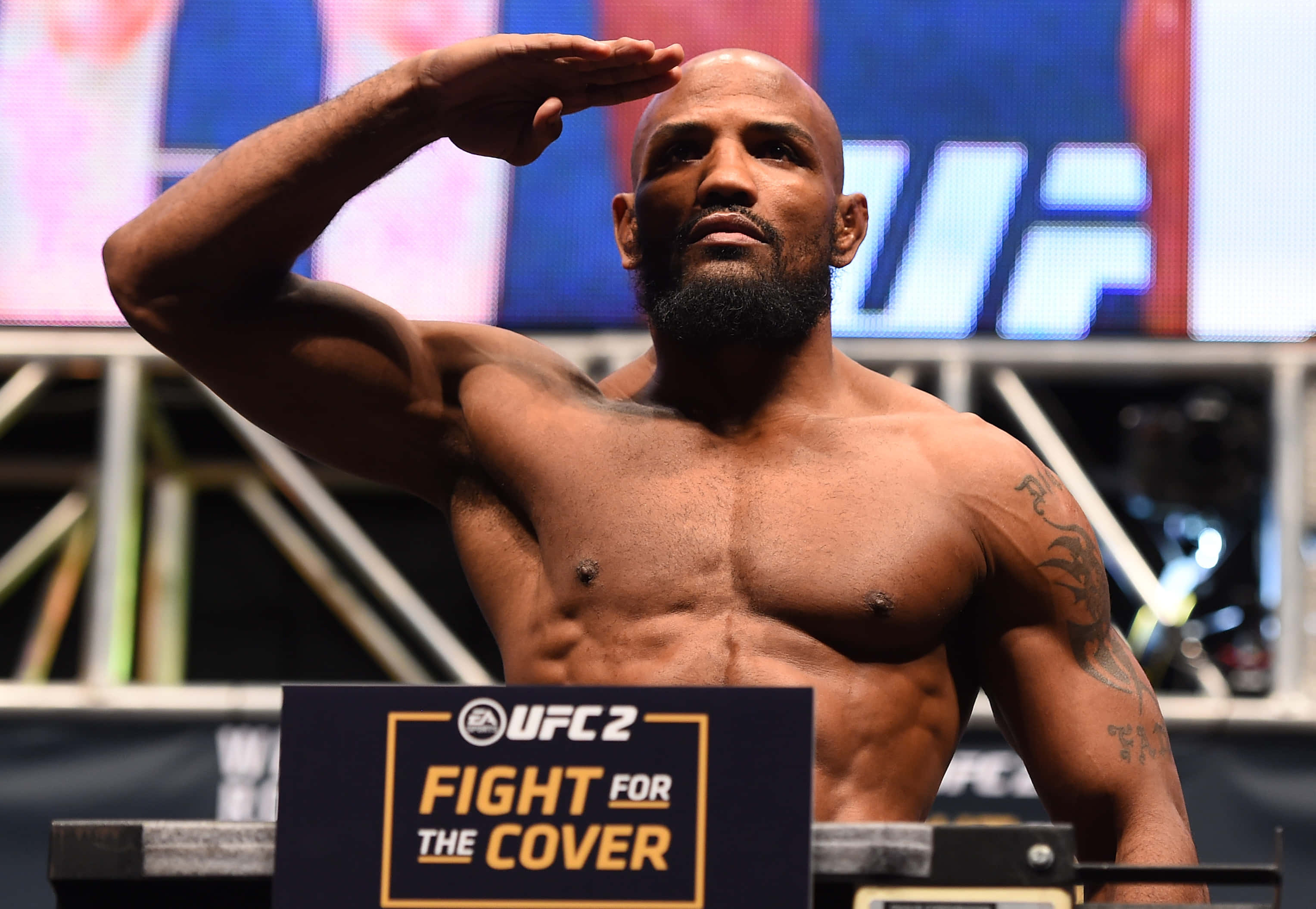 約爾·羅梅羅 UFC 194 称重儀式 壁紙 — 一位肌肉發達、光頭、留鬍子的戰士在明亮的競技場燈光下，自信地在 UFC 稱重活動中敬禮。