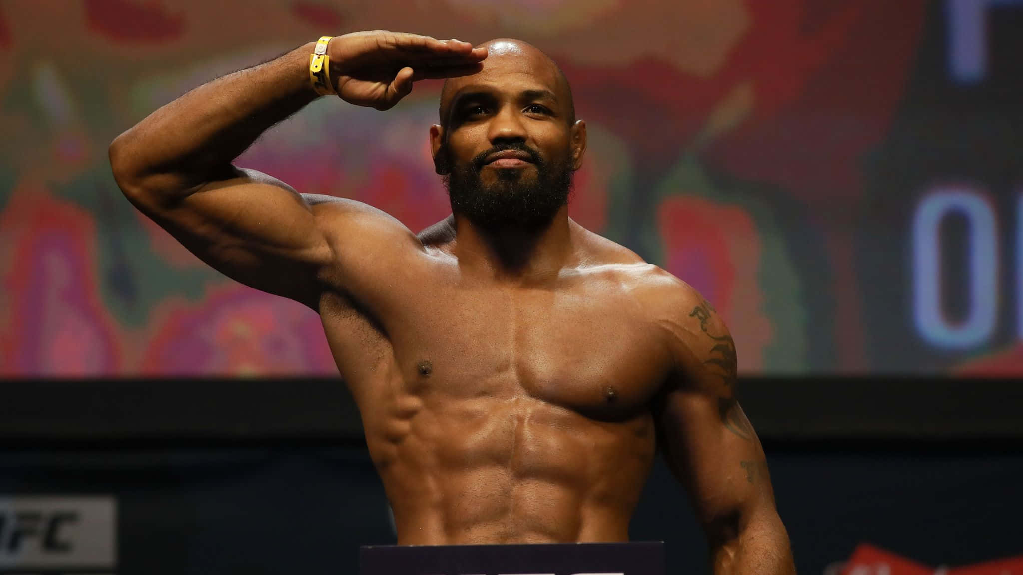 約爾·羅梅羅 UFC 205體重確認儀式 壁紙 — 一位肌肉發達、光頭、留鬍子的戰士，在色彩繽紛的模糊背景下，自信地在UFC稱重中敬禮。