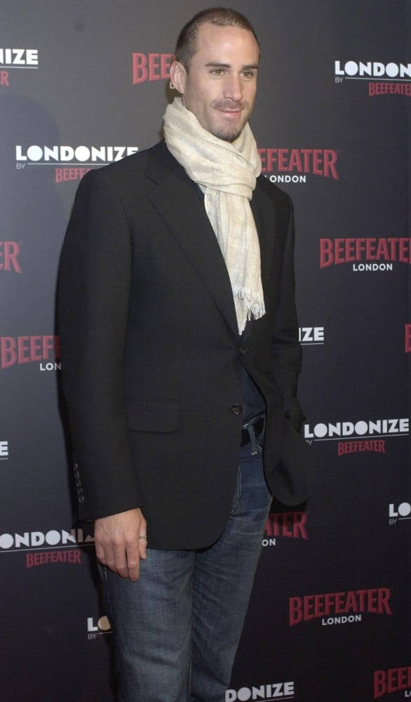 約瑟夫·費尼恩出席Beefeater活動 壁紙 — 一名身穿黑色西裝外套和奶油色圍巾的男子，在Beefeater London活動中自信地站在品牌背景前。