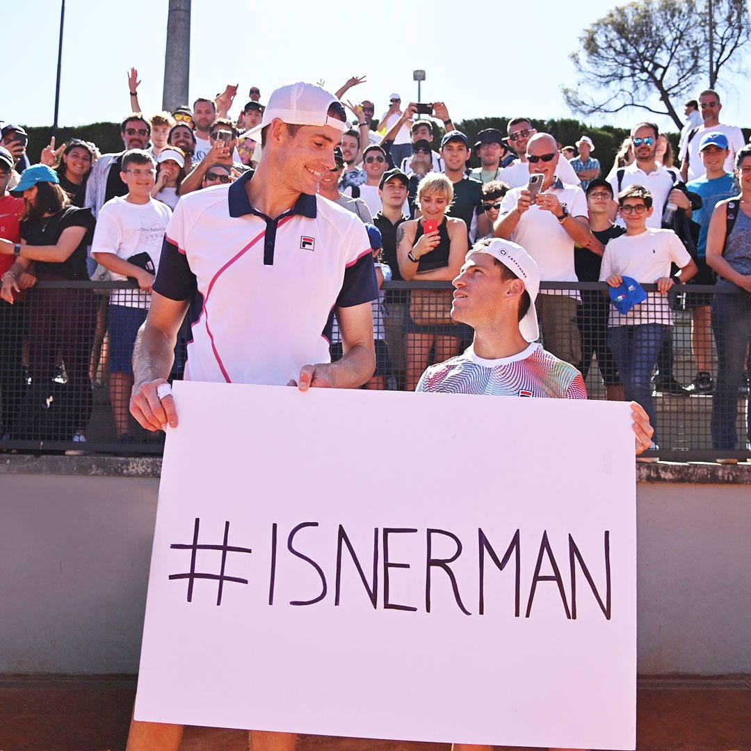 兩位網球選手舉著 #ISNERMAN 標語牌，身後有歡呼的粉絲。