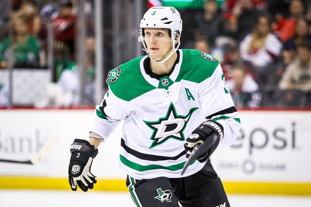 約翰·克林格伯格 達拉斯星隊 NHL 球員 壁紙 — 一名達拉斯星隊的冰球運動員穿著綠色和白色球衣，在比賽中於冰上激烈滑行，背景有觀眾。