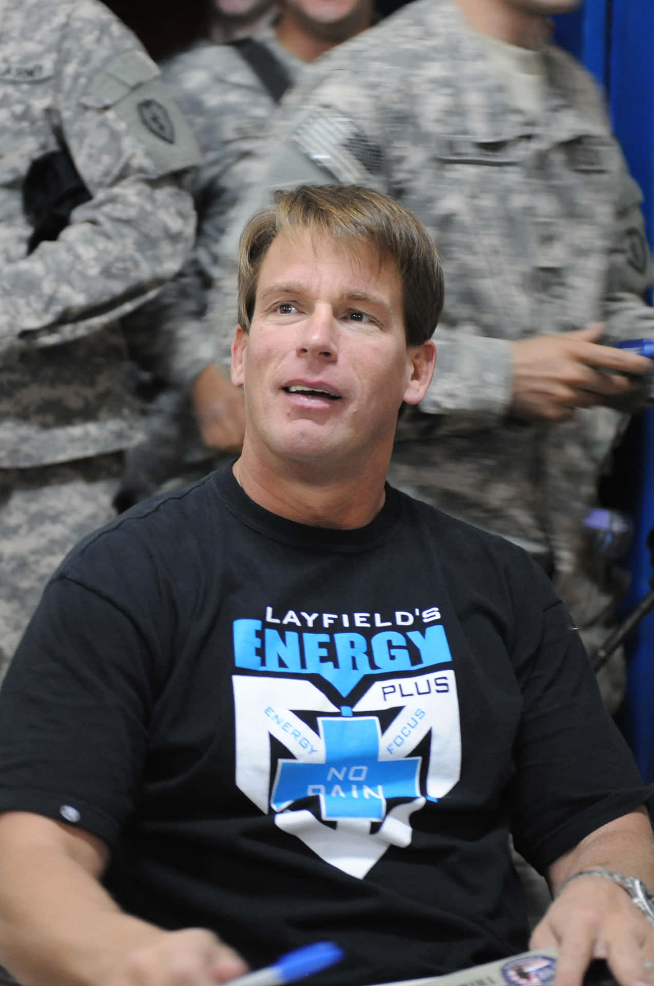 約翰·布拉德肖·萊夫爾與士兵互動 壁紙 — 一名身穿印有「Layfield's Energy Plus」圖案黑色T恤的男子，坐在模糊的迷彩軍人前面。