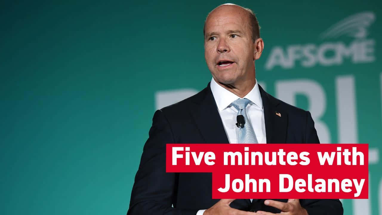穿西裝的男子在活動中發言，橫幅寫着 'Five minutes with John Delaney'。