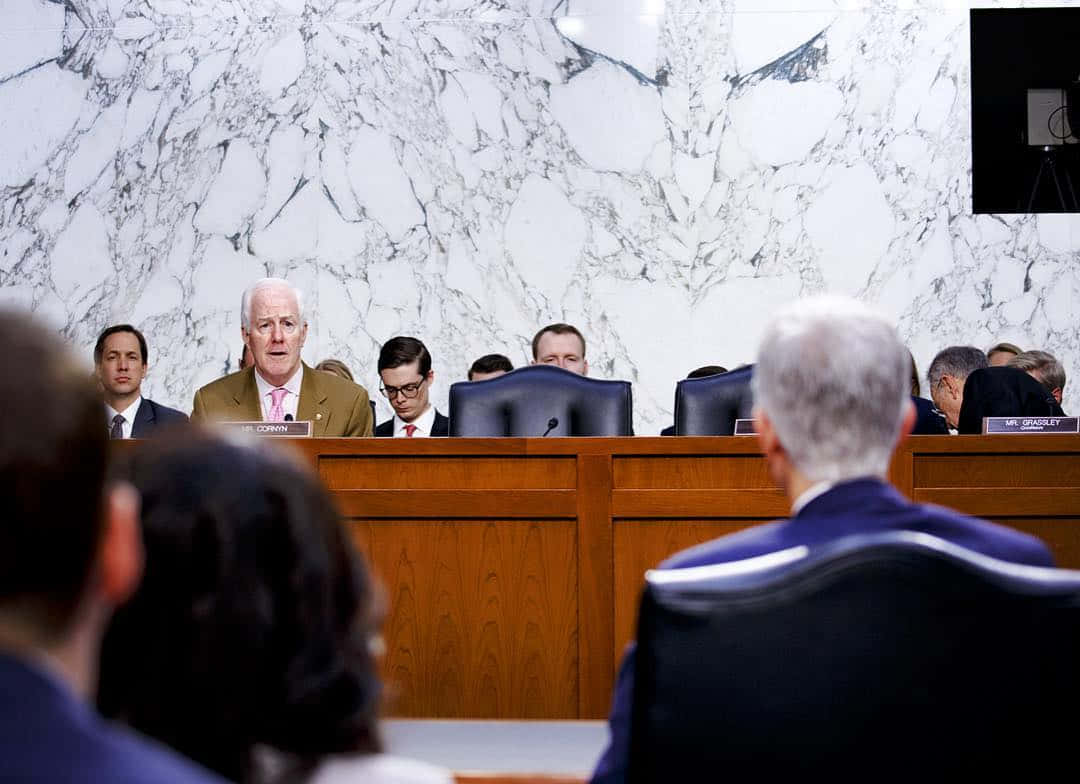 一名身穿米色西裝的男子在政府聽證會上發言，其他人聆聽。