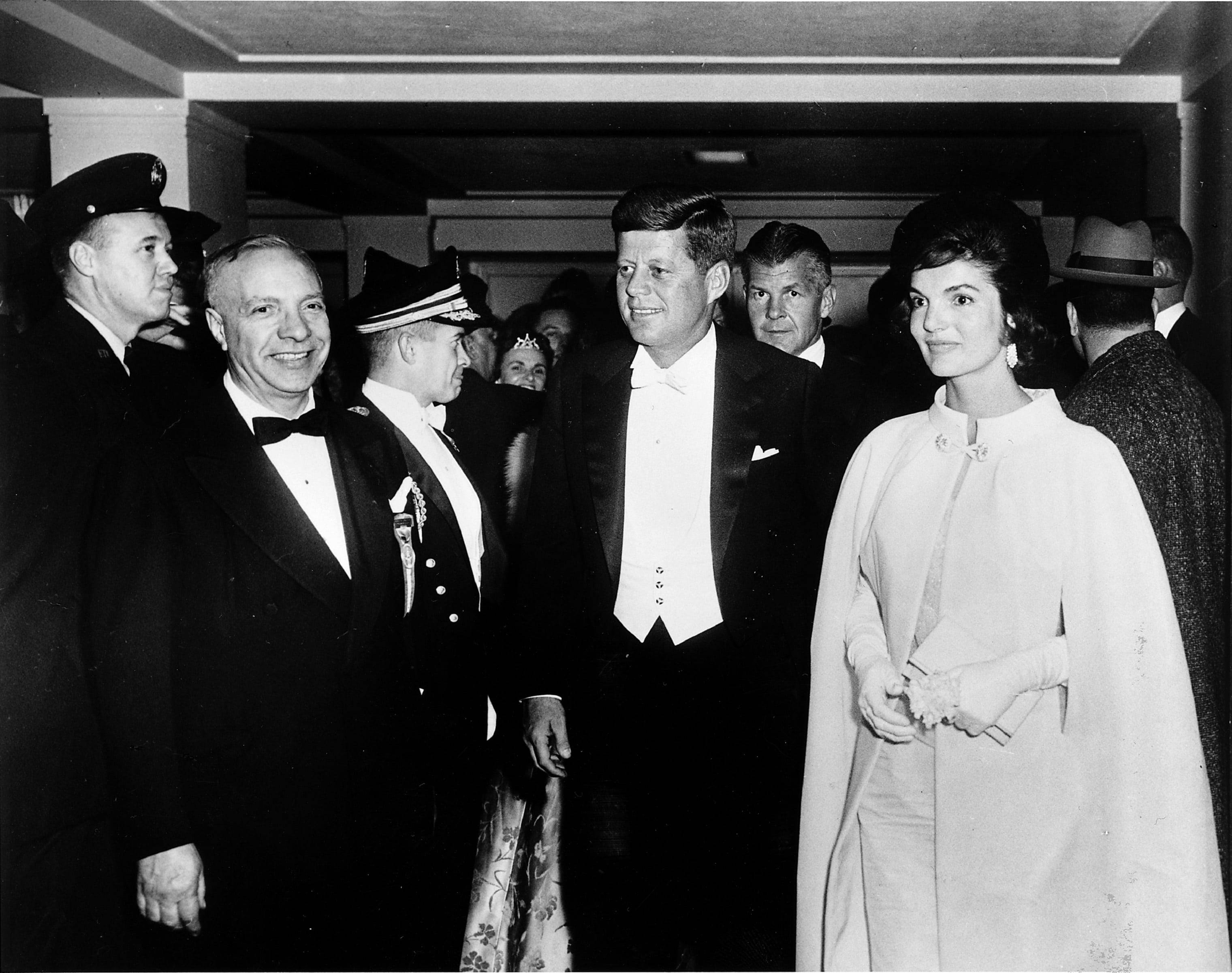 約翰·肯尼迪與妻子傑奎琳 壁紙 — JFK 與積琪蓮·甘迺迪在正式室內聚會中微笑的黑白照片，周圍有穿制服的官員和賓客。