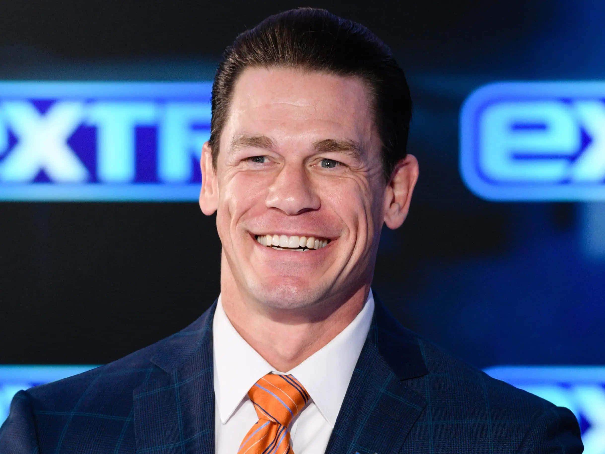John Cena 桌布 壁紙 — 約翰·辛納在閃耀的藍色「Extreme」背景下，穿著鋒利的藍色格紋西裝和橙色領帶，燦爛地微笑。