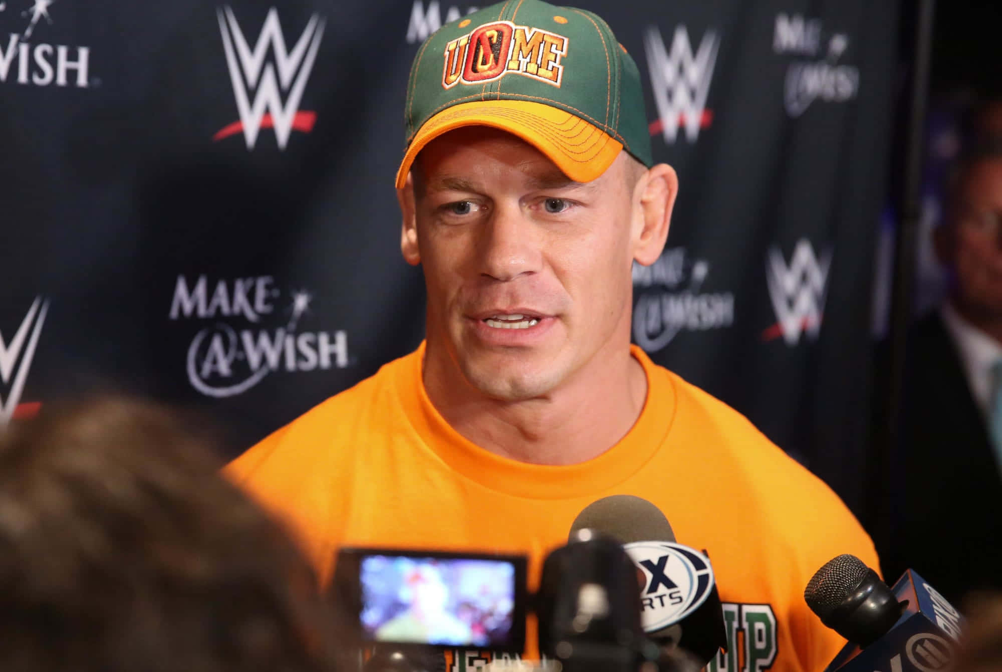 John Cena 桌布 壁紙 — 約翰·辛納身穿橙色襯衫和綠色帽子，在WWE Make-A-Wish活動中對著麥克風向記者講話。