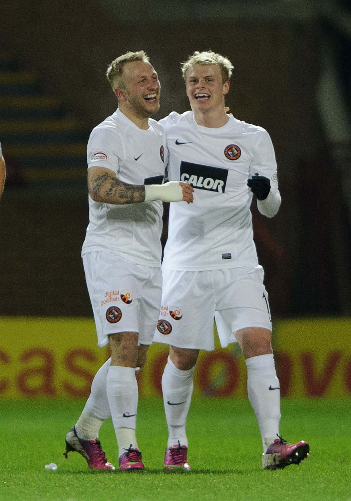 約翰尼·羅素 邁克爾·鄧迪聯足球俱樂部 壁紙 — 兩名身穿白色球衣的快樂足球員在體育場燈光下，於充滿活力的綠色球場上慶祝入球。