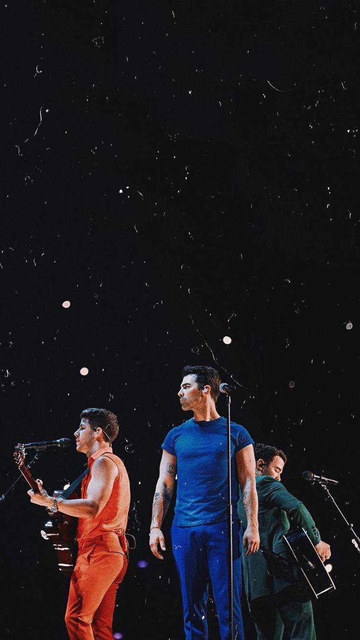 約翰斯兄弟舞台演唱 壁紙 — 三位音樂家在星光燈光下表演，周圍有五彩紙屑飄落。
