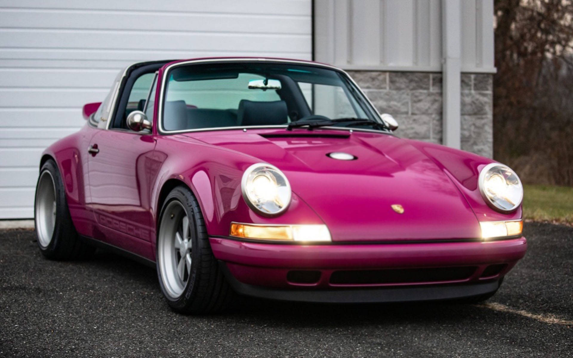 紅寶石紅Singer Porsche 壁紙 — 一輛亮面洋紅色保時捷911 Targa停在白色車庫門前的瀝青路上，車頭燈亮著。