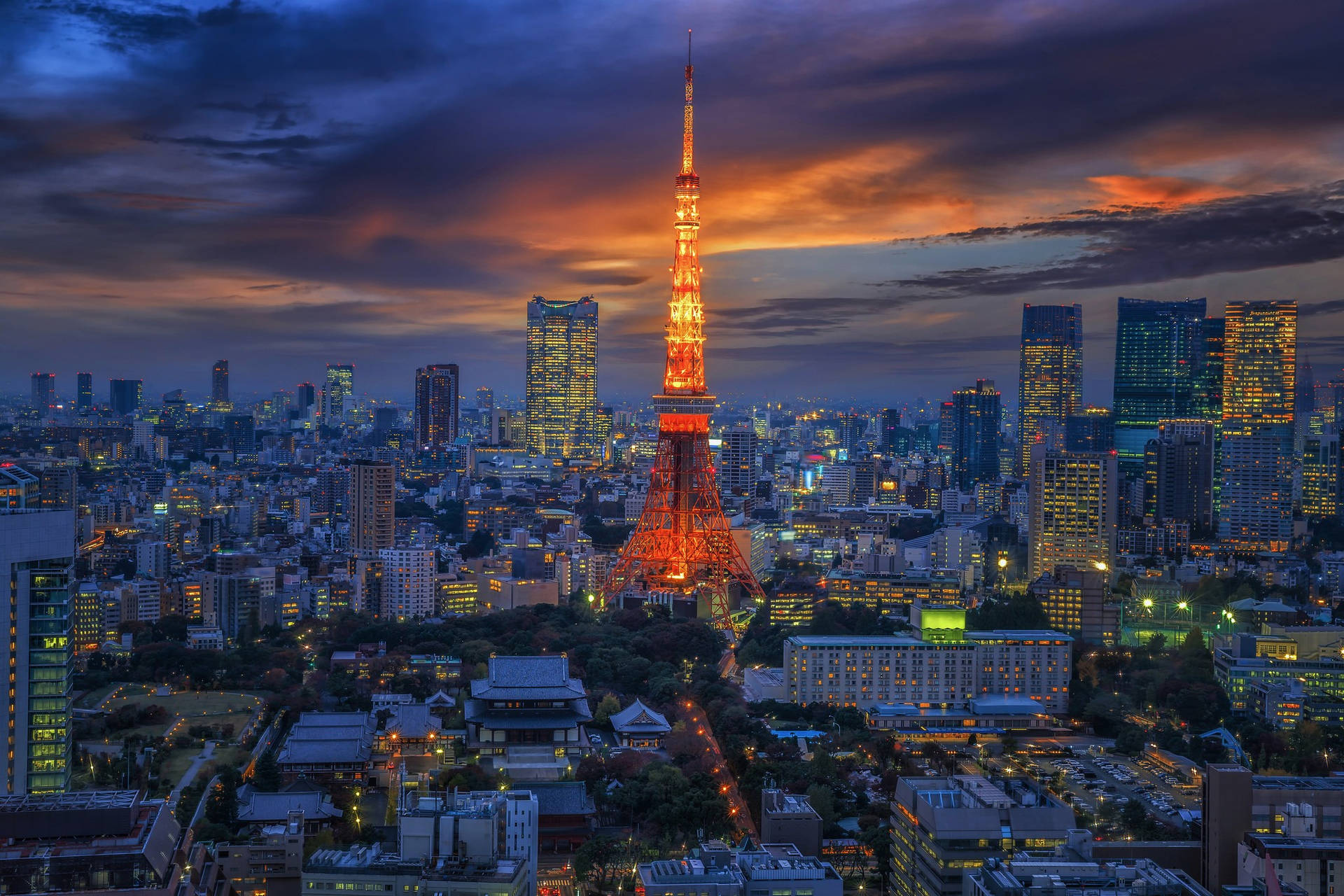紅橙色東京塔 壁紙 — 東京鐵塔在戲劇性的暮色天空中溫暖地發光，城市燈光照亮了下面的城市擴展區。