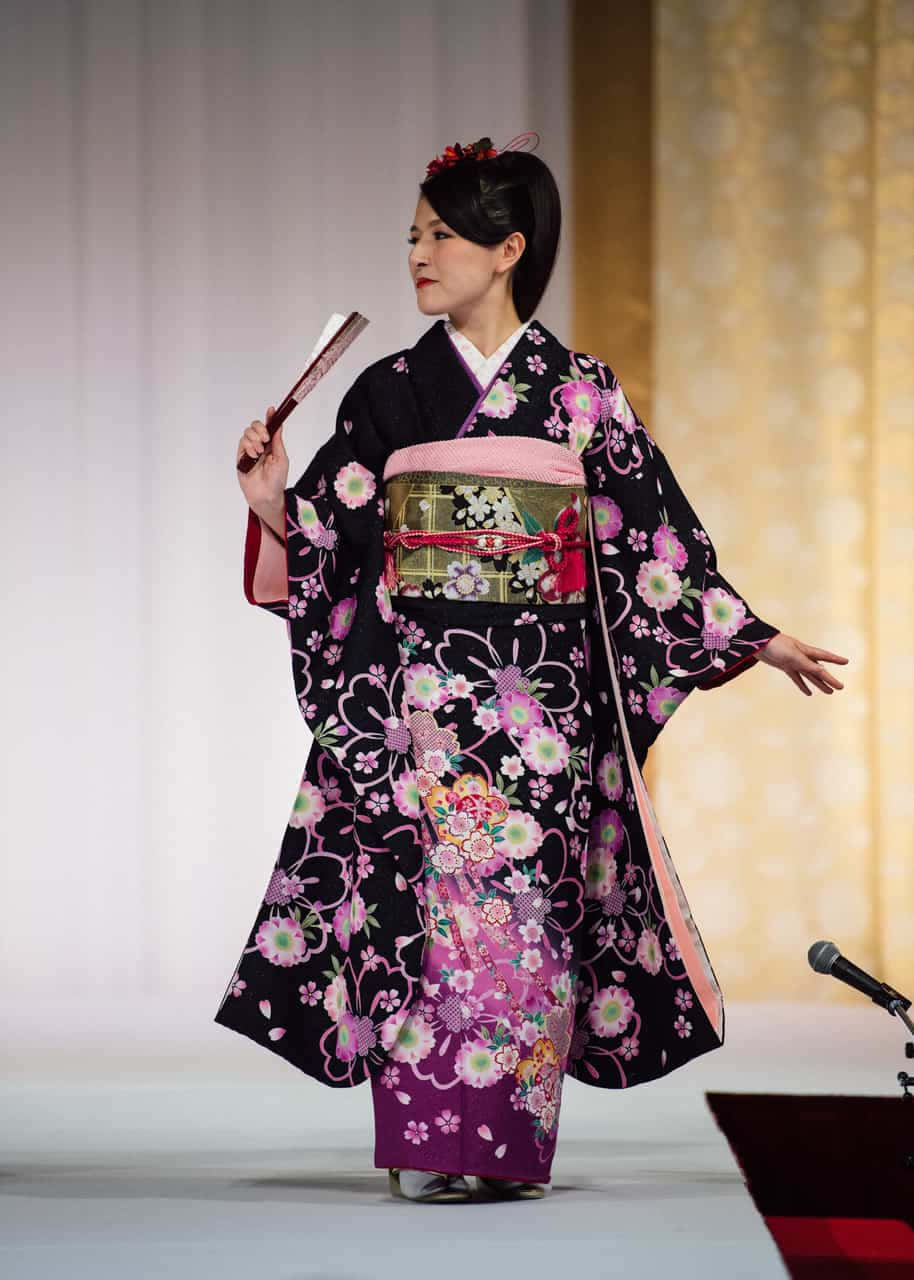 紅橙色調的傳統日本和服 壁紙 — 一位身穿黑粉花卉和服的女士優雅地在舞台上擺姿勢，手持扇子。