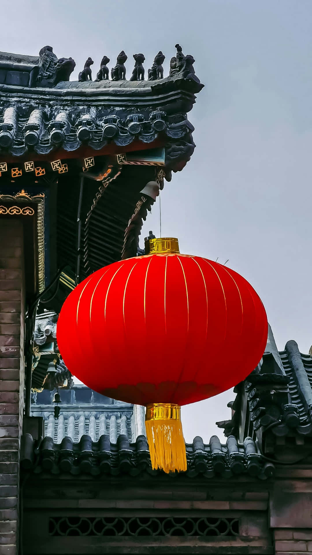 紅燈籠懸掛於傳統屋簷 壁紙 — 一個鮮紅色的燈籠帶有金色流蘇，在陰天背景下懸掛在傳統中式瓦屋頂前，格外顯眼。