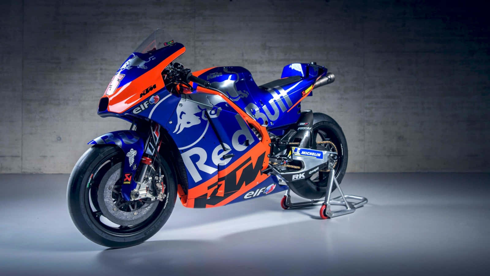 紅牛 KTM 圖片 壁紙 — 一架帶有Red Bull品牌標誌的時尚藍橙色KTM MotoGP賽車，優雅地停在簡約混凝土背景前的支架上。