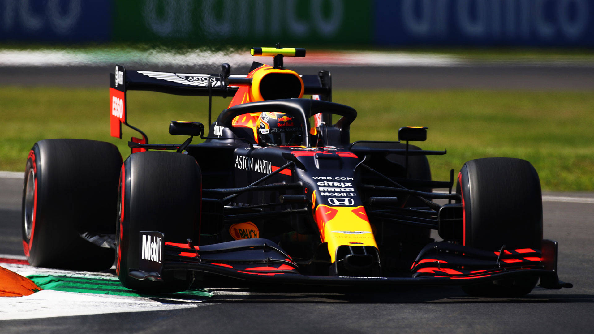 紅牛隊亞歷山大·阿爾本賽車 壁紙 — 一架帶有Aston Martin品牌標誌的Red Bull Racing一級方程式賽車，在明亮的日光下沿著賽道飛馳，賽道兩旁是綠色草地和橙色路緣石。