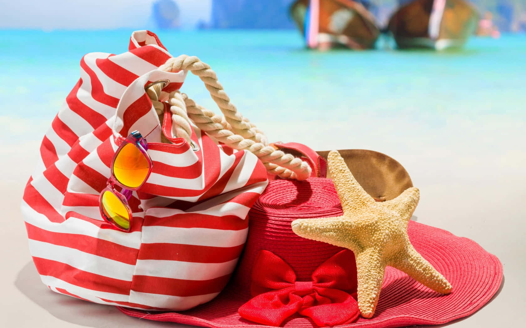 Beach Bag 桌布 壁紙 — 紅白條紋沙灘袋配太陽眼鏡，旁邊放著一頂飾有海星的紅色太陽帽，放在靠近碧綠海水的柔軟沙灘上。