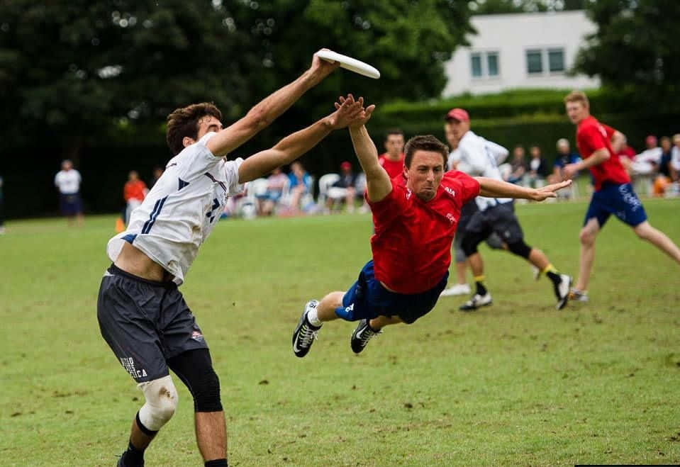 紅白隊伍最佳 Ultimate Frisbee 背景 壁紙 — 兩名男子在草地上跳躍並伸手接住飛行的飛盤，背景有觀眾。