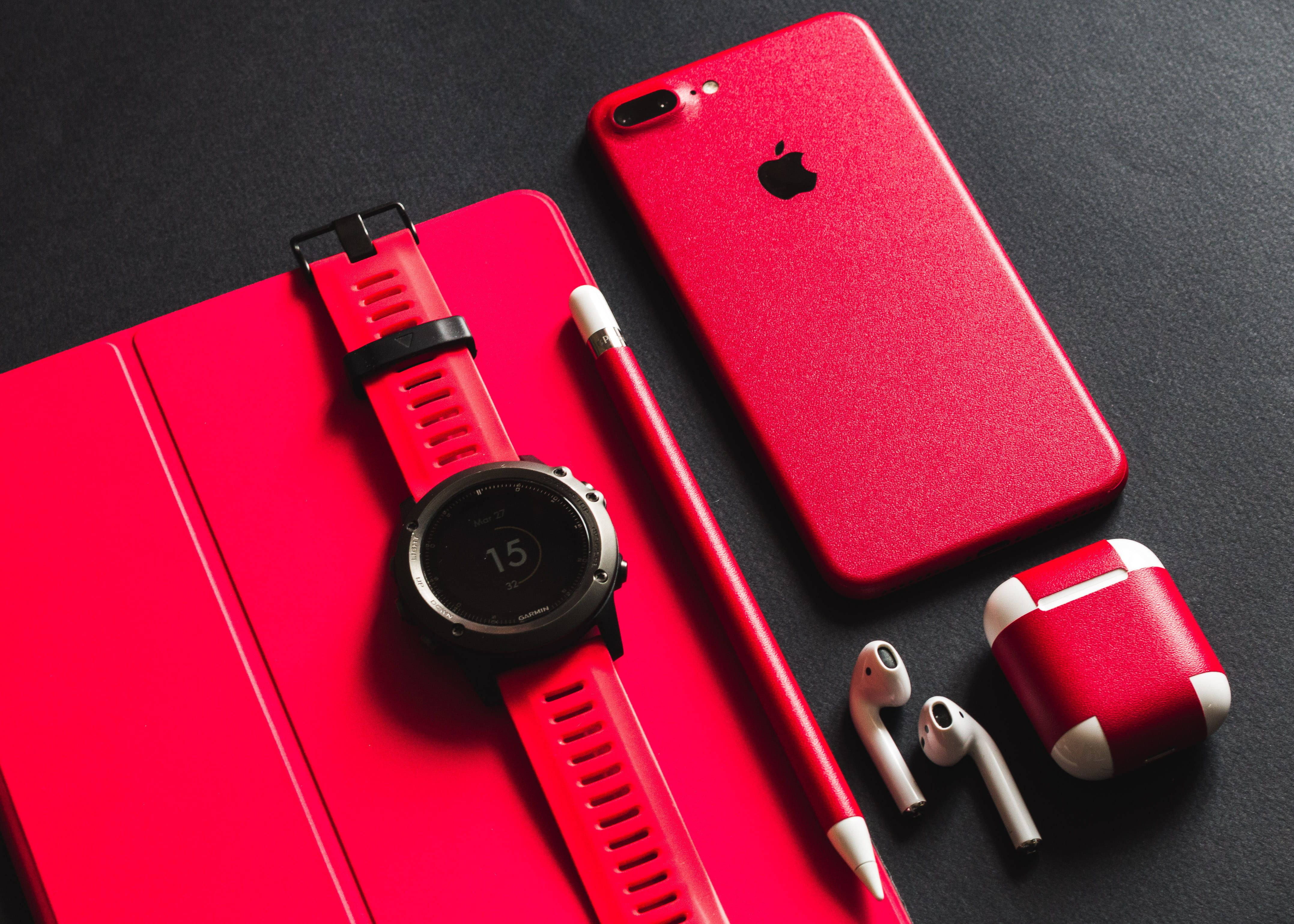 紅色 Apple AirPods 壁紙 — 一個大膽的紅色科技設置，包括智能手錶、電話、耳機和觸控筆，排列在深色表面上。