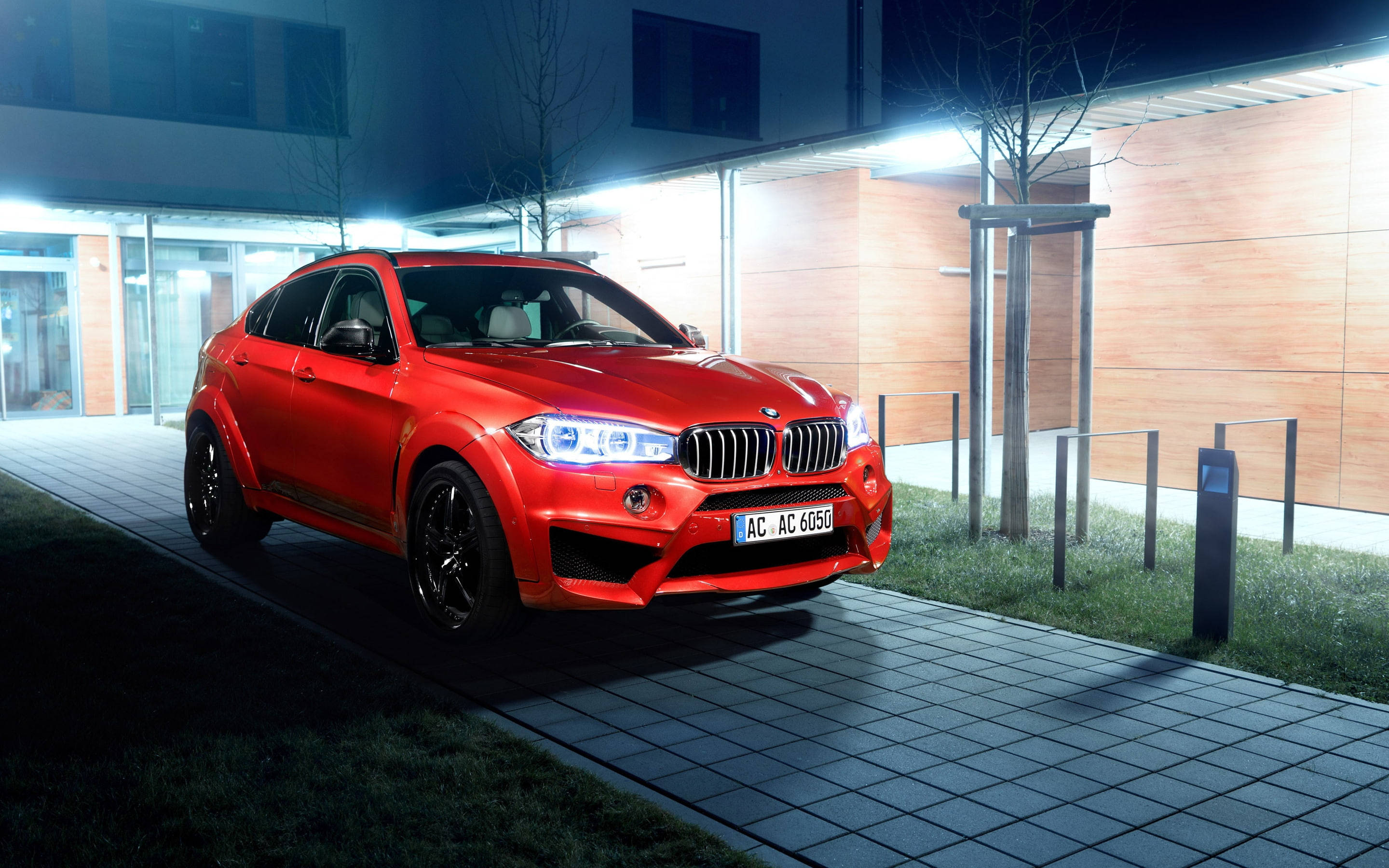 紅色BMW X6 M Schnitzer Falcon閃耀於屋外 壁紙 — 一輛鮮紅色的BMW X6M AC Schnitzer在夜間停泊在現代建築旁邊的瓷磚行人路上，建築物有發光的燈光。