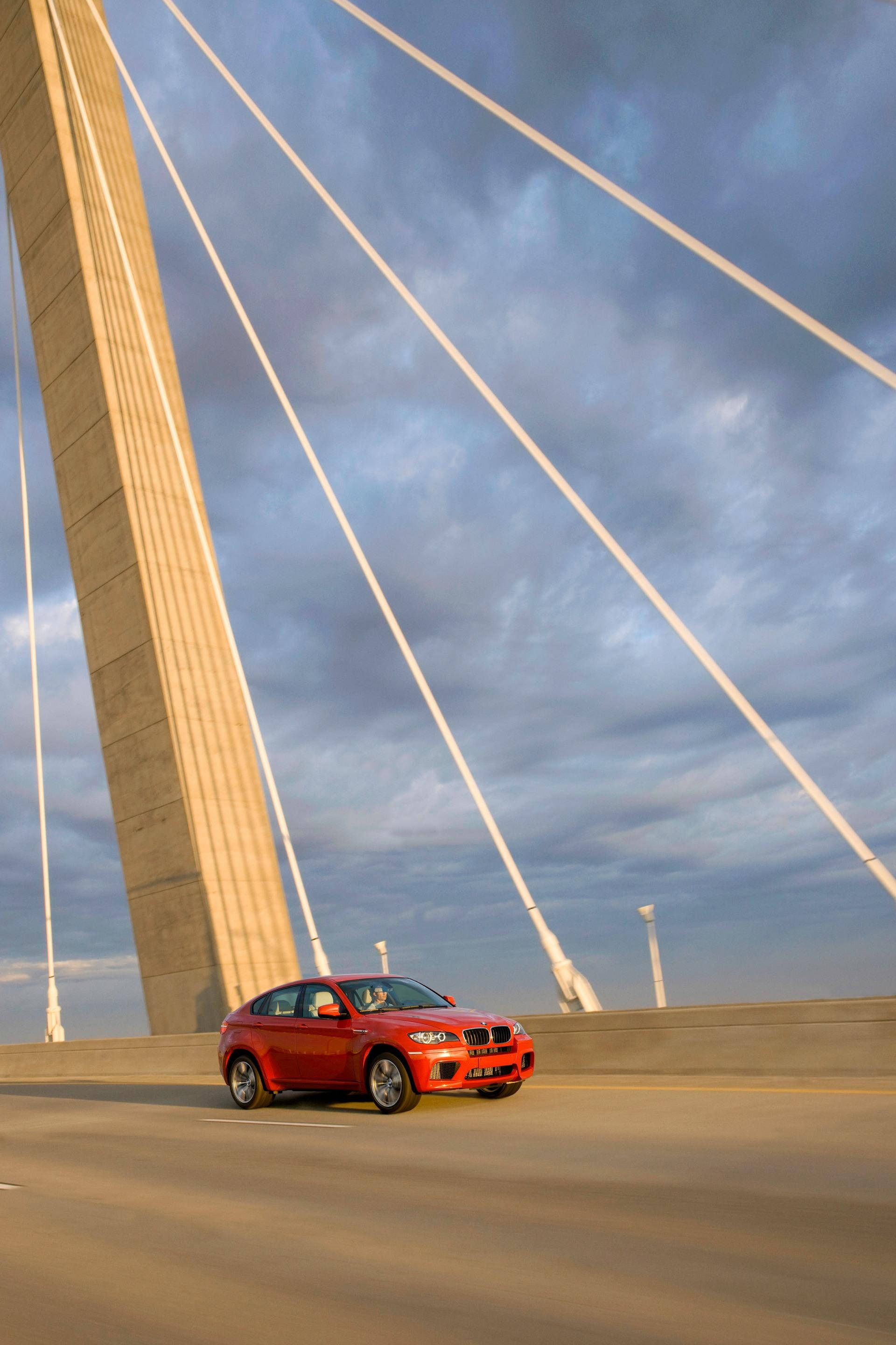 紅色BMW X6 M行駛於橋上 壁紙 — 一輛鮮紅色的BMW X6在戲劇性的多雲天空下行駛於現代斜拉橋上。