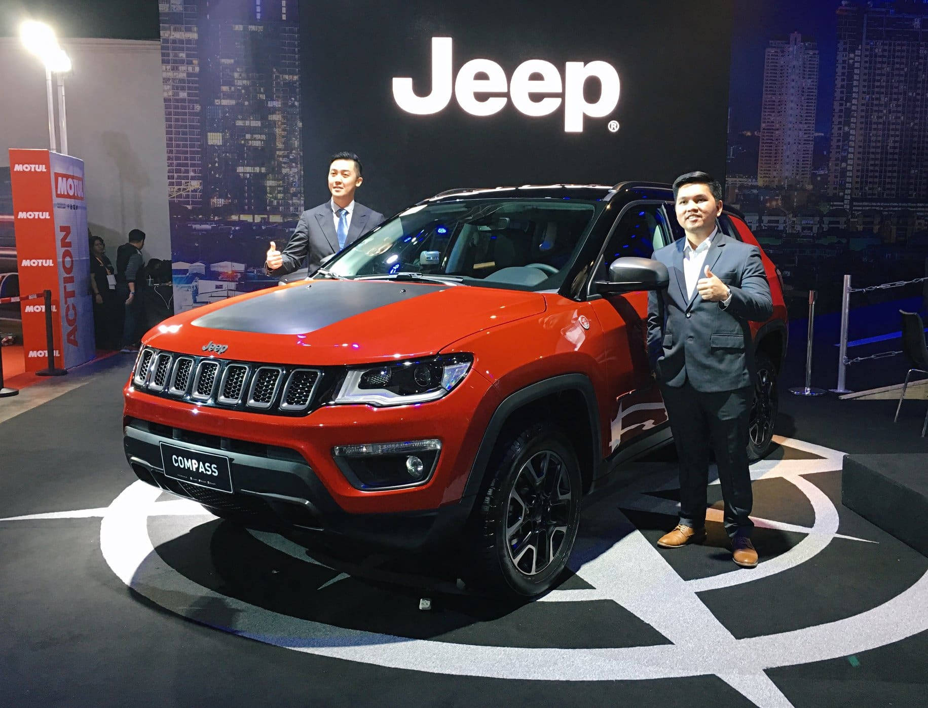 兩名穿西裝的男子在車展上於一輛紅色Jeep Compass旁邊豎起大拇指。