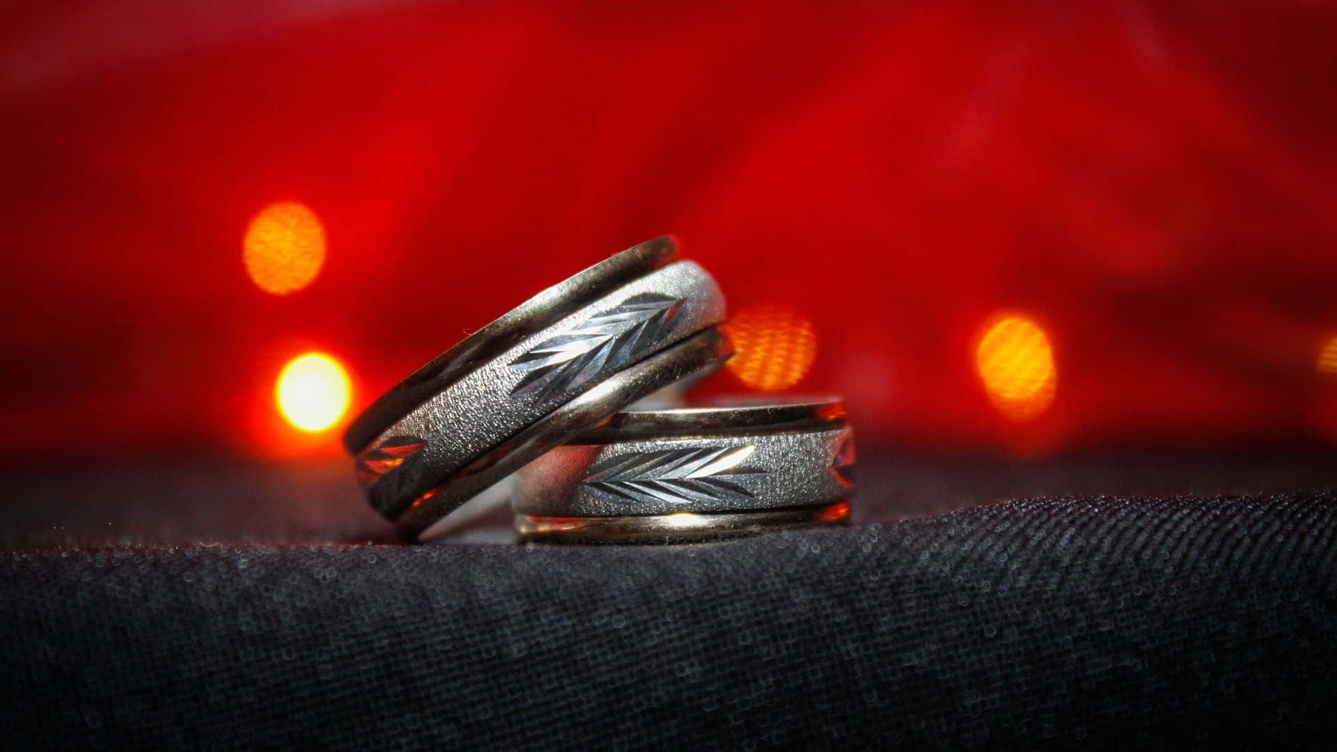 兩枚雕刻的銀色結婚戒指在深色布料上，背景有紅光。