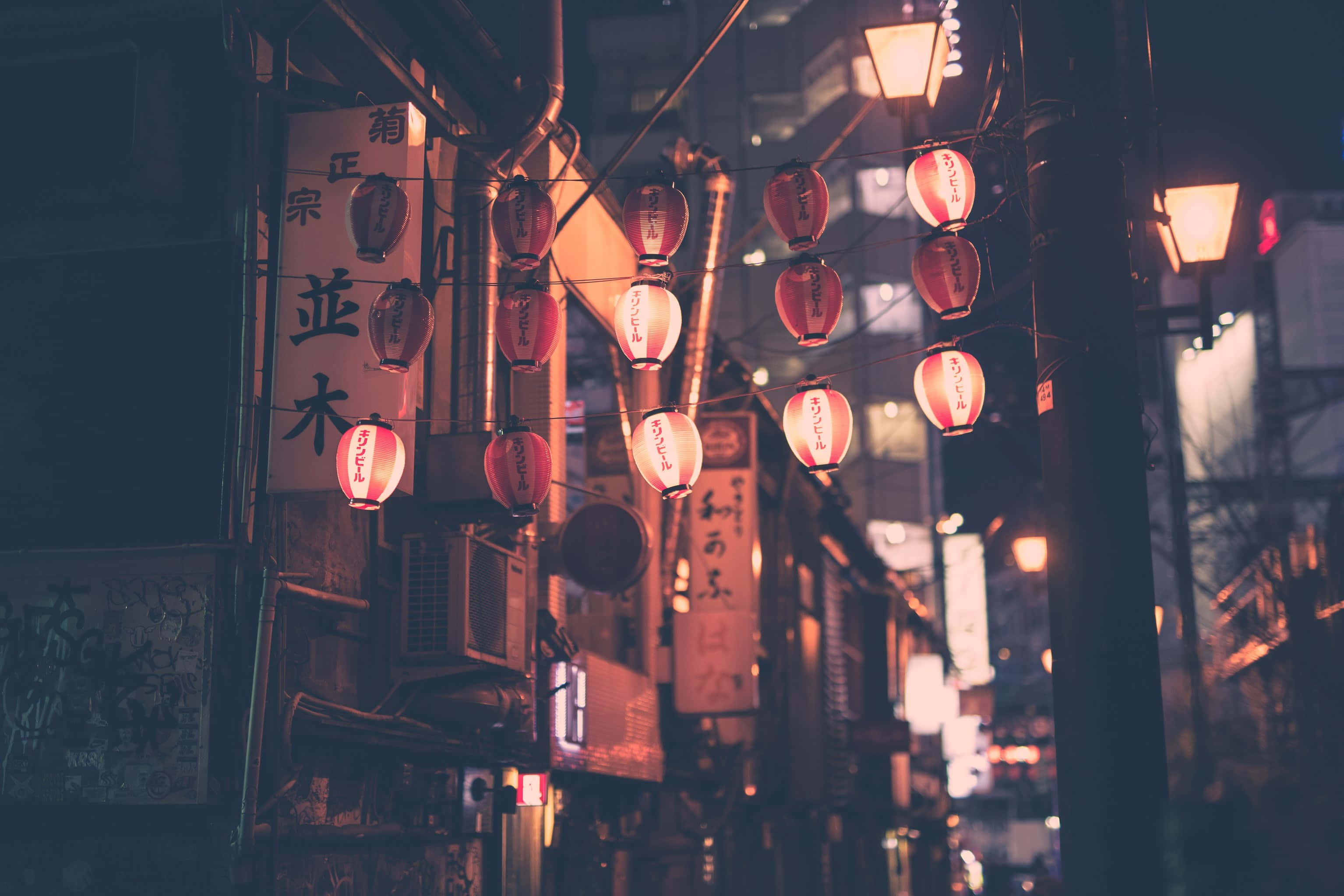 // 紅色日本扇子揮手問好 壁紙 — 溫暖發光的紅色燈籠掛在狹窄的日本小巷中，夜晚時分，柔和的光線灑在風化的建築物和招牌上。