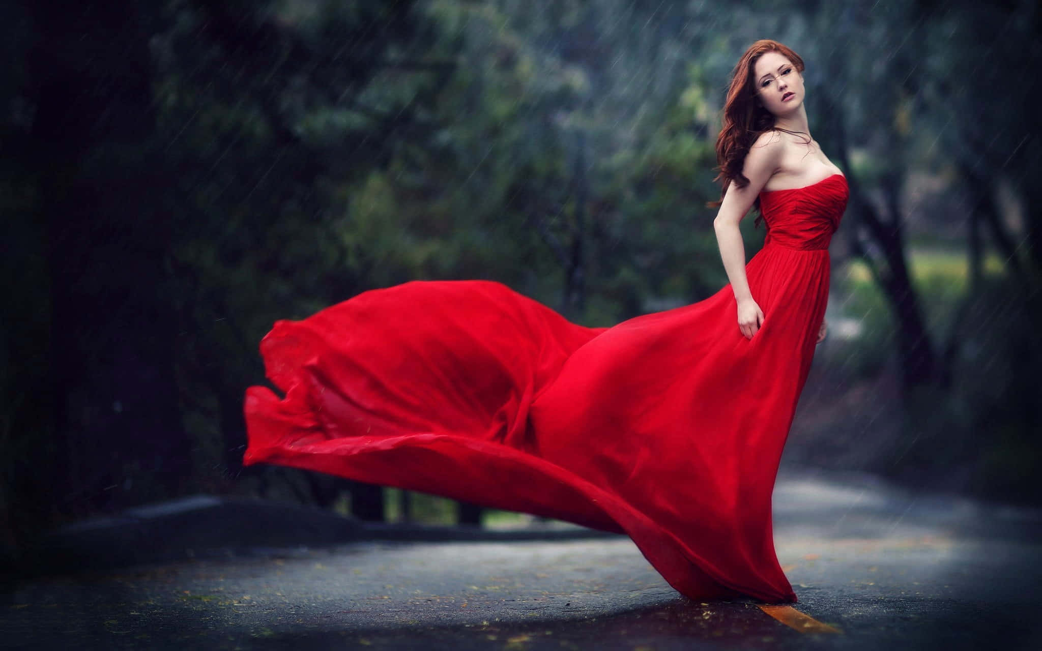 紅色時尚禮服圖片 壁紙 — 一位身穿飄逸紅裙的女子站在被雨水浸濕的森林小徑上，裙擺在風中戲劇性地飄揚。