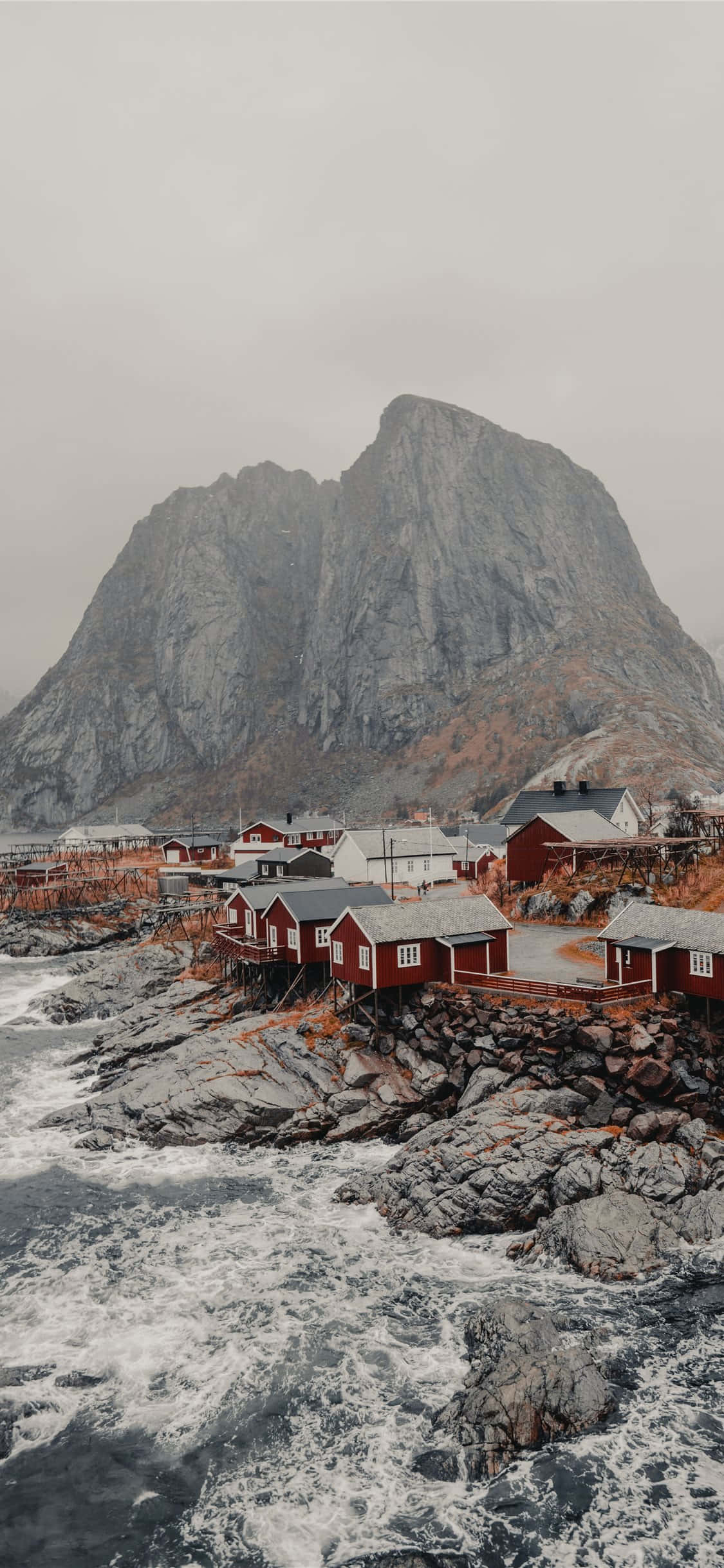 Norway 桌布 壁紙 — 紅色木屋依附在岩石海岸上，聳立著灰色的山脈，天空多雲。